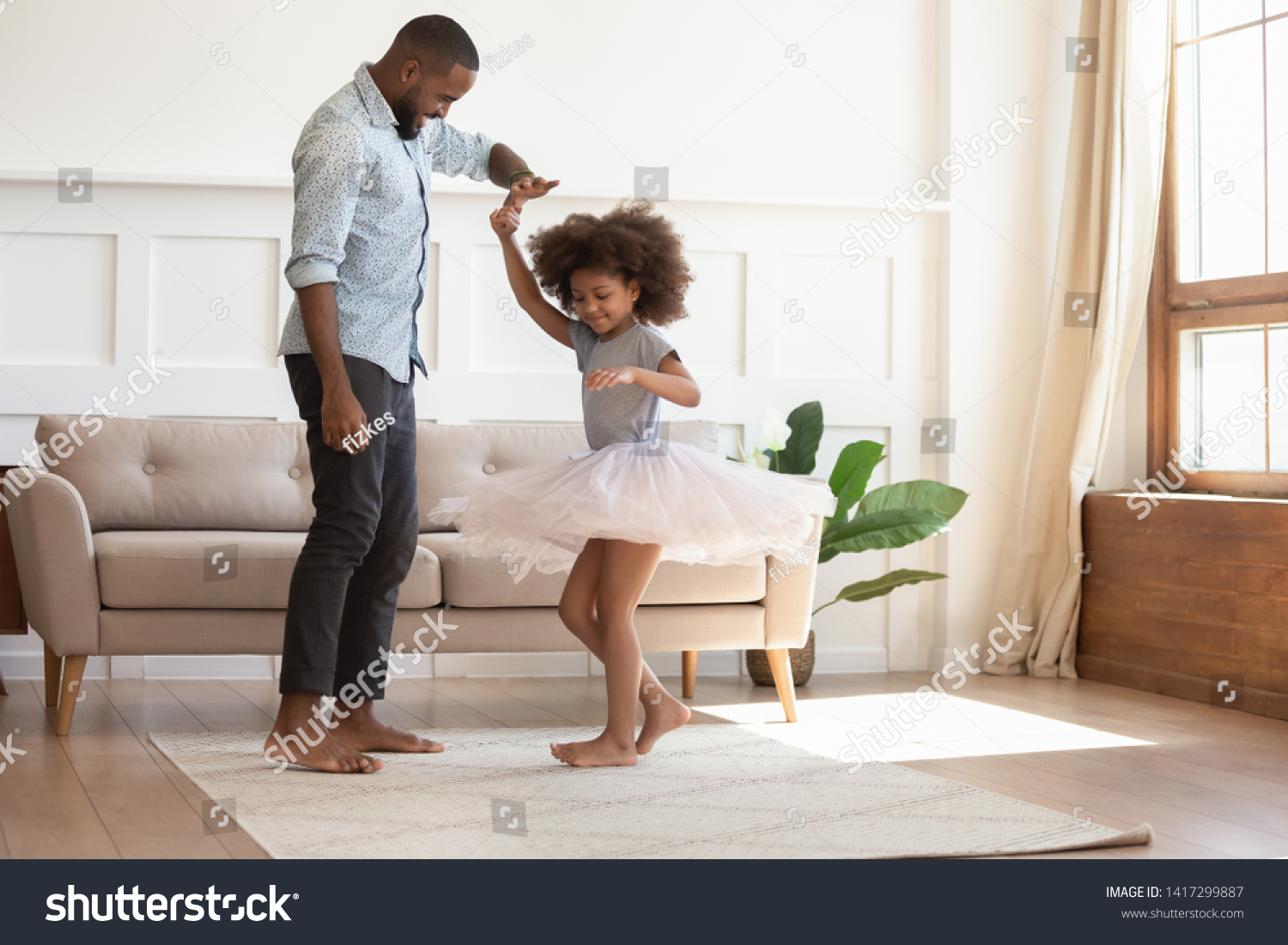 Loving black dad holding hand of child daughter dancing with his child princess wearing skirt, happy young african father and little cute daughter playing in living room enjoy time together at home #1417299887