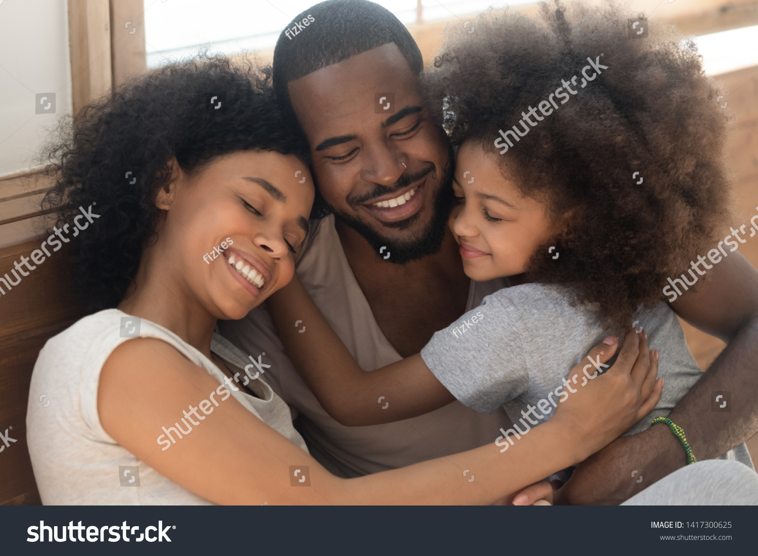 Happy affectionate african american family of three bonding embracing, cute kid child daughter and loving parents cuddling congratulating dad with fathers day hugging smiling daddy laughing together #1417300625
