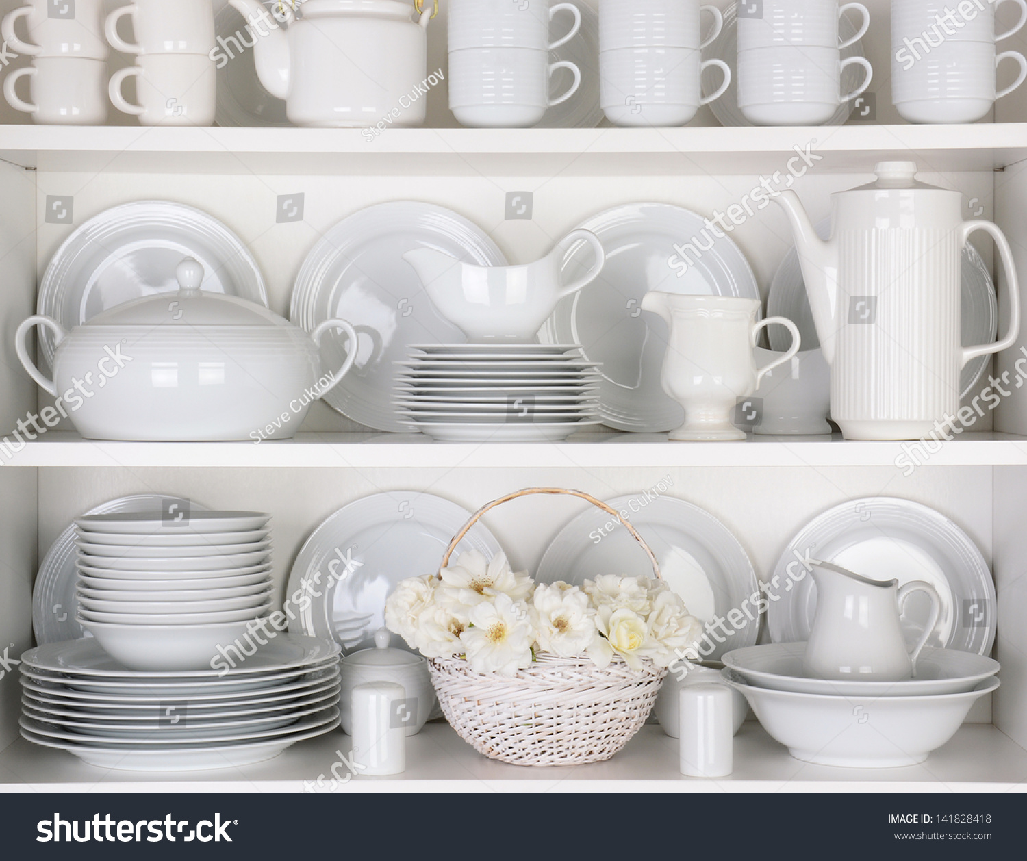 Closeup of white plates and dinnerware in a cupboard. A basket of white roses is centered on the bottom shelf. Items include, plates, coffee cups, saucers, soup tureen, tea pot, and gravy boats. #141828418