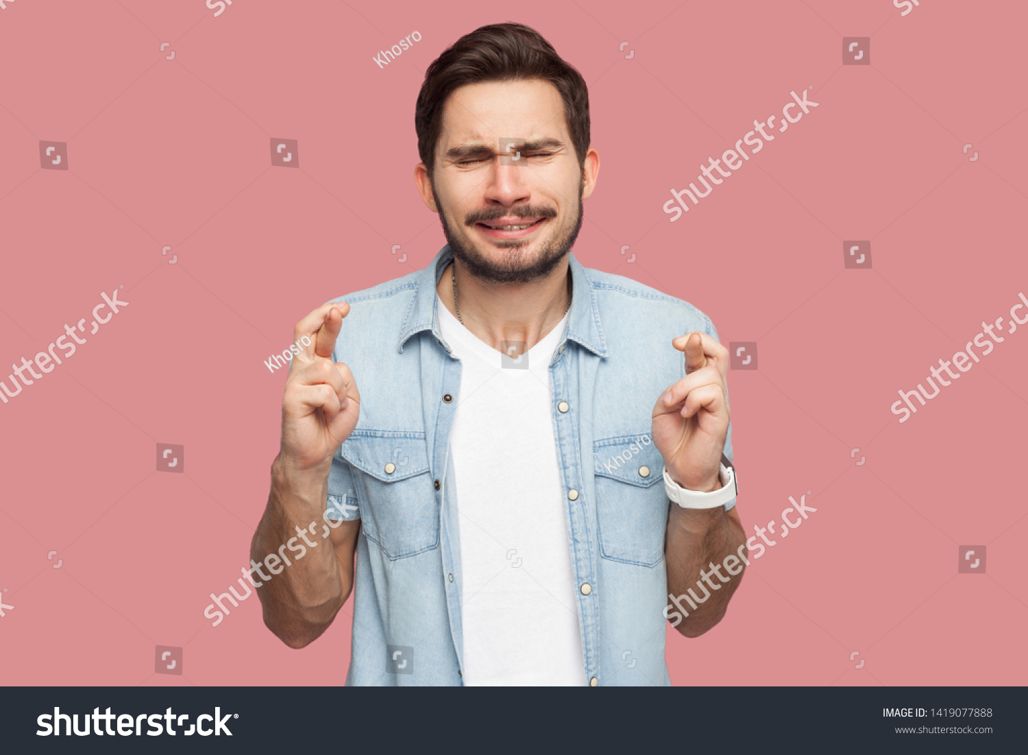 Portrait of hopeful handsome bearded young man in blue casual style shirt standing with crossed fingers, closed eyes and hope to win. indoor studio shot, isolated on pink background. #1419077888