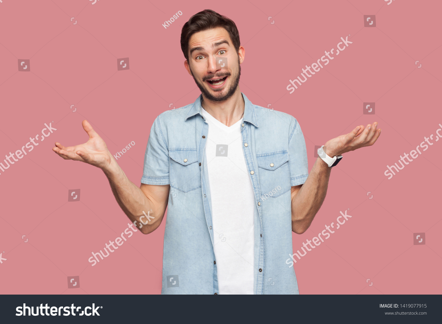 Portrait of surprised handsome bearded young man in blue casual style shirt standing with raised arms and looking at camera with amazed face. indoor studio shot, isolated on pink background. #1419077915