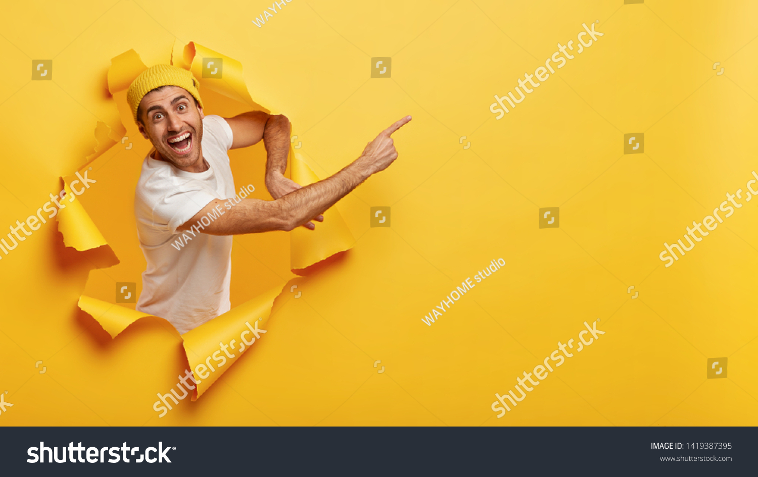 Confident happy man in casual white t shirt, points at upper right corner, invites going in this direction, demonstrates welcoming gesture, stands in torn paper hole, says you must see product #1419387395