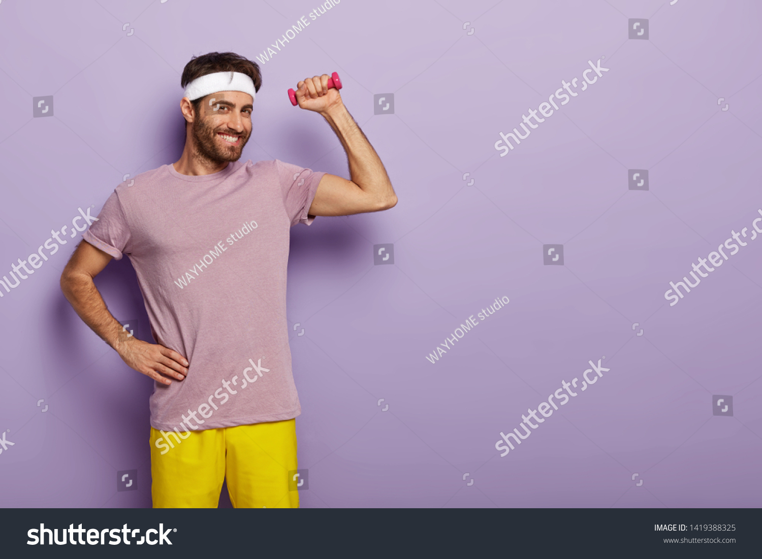 Glad smiling male gymnast keeps one hand on waist, raises muscualr arm with dumbbell, satisfied after having training in gym, dressed in casual t shirt and shorts isolated over purple wall empty space #1419388325