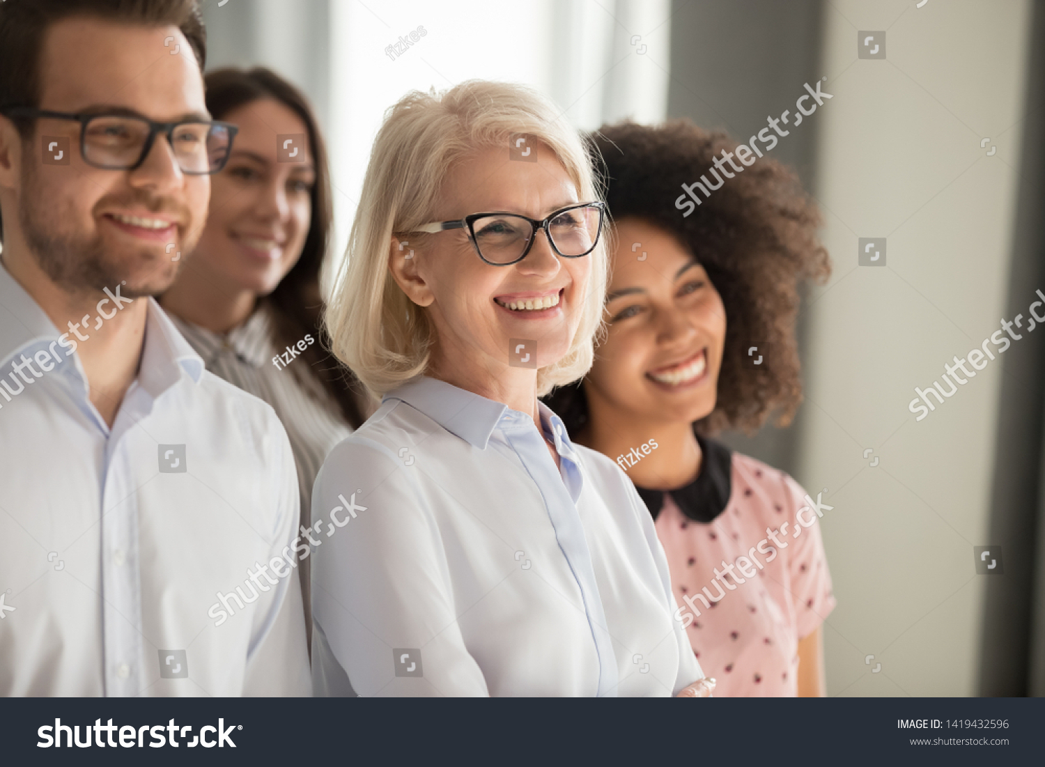 Side view of smiling diverse employees stand in row together posing for group picture in office, happy confident multiethnic team look at camera making photo, showing unity. Teamwork concept #1419432596
