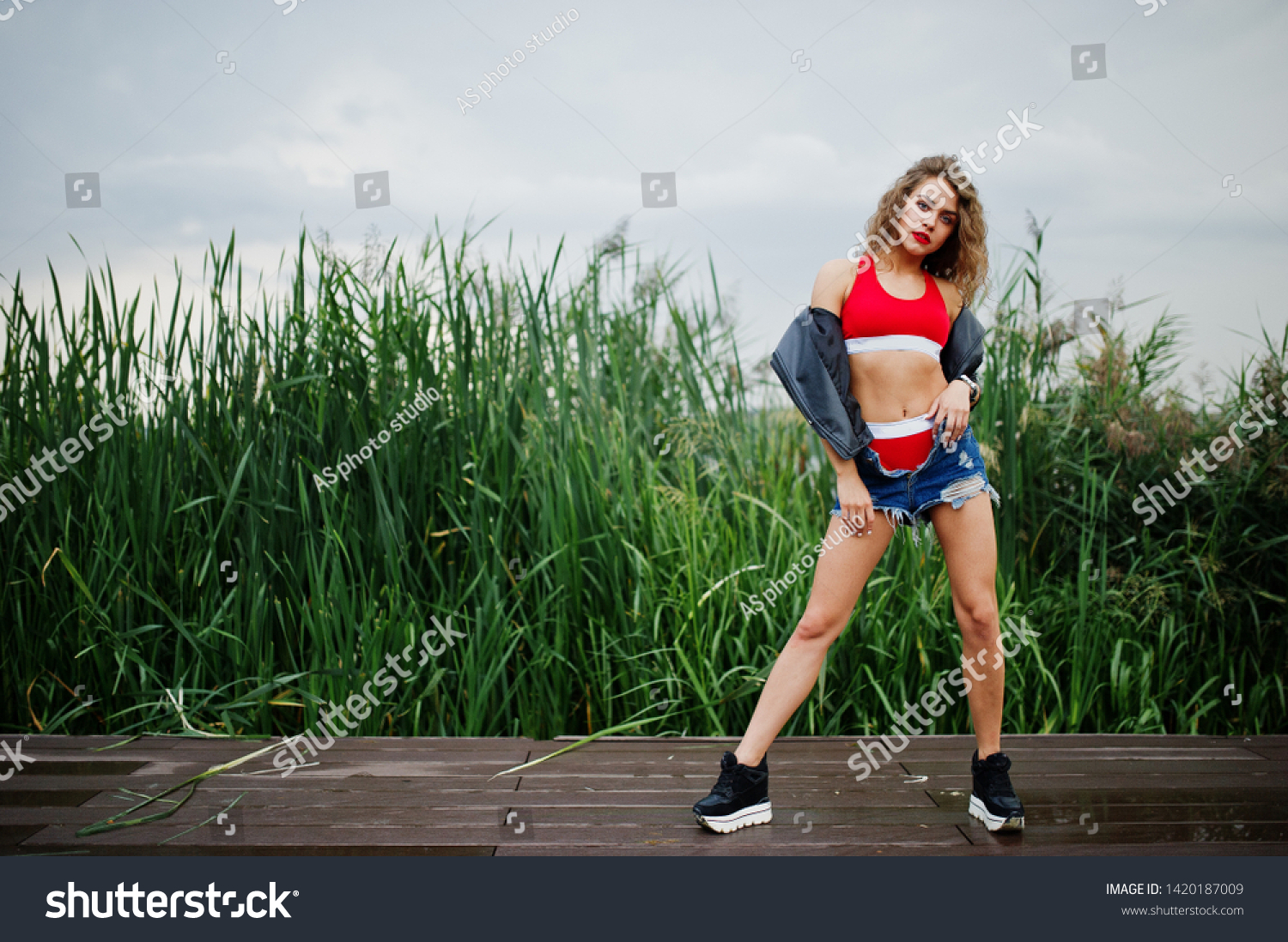 Sexy curly model girl in red top, jeans denim shorts, leather jacket and sneakers posed in the reed. #1420187009