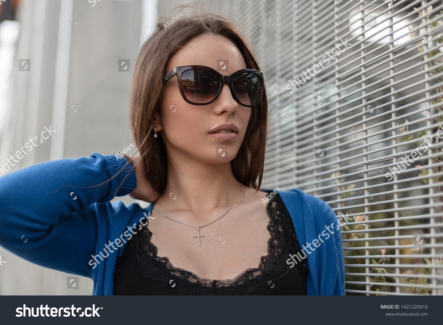 Pretty attractive young woman hipster with sexy lips in stylish sunglasses in a stylish blue knitted cape in a fashionable black top with lace and posing near of a modern metal wall. Beautiful girl. #1421220416