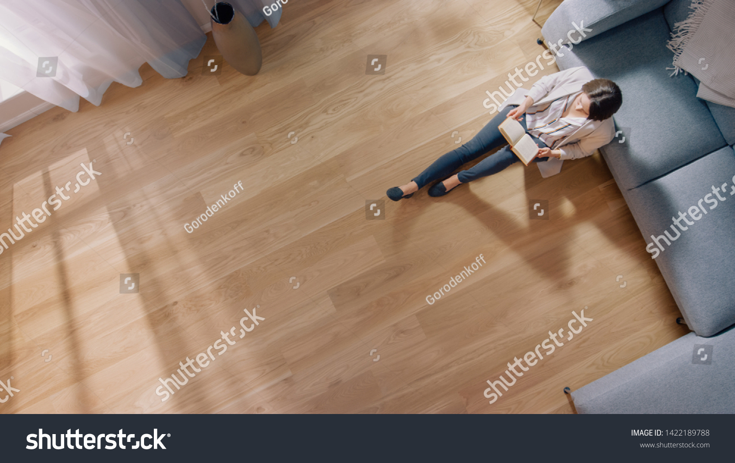 Young Woman is Sitting on a Floor and Reading a Book. Cozy Living Room with Modern Interior, Grey Sofa and Wooden Flooring. Top View Camera Shot. #1422189788
