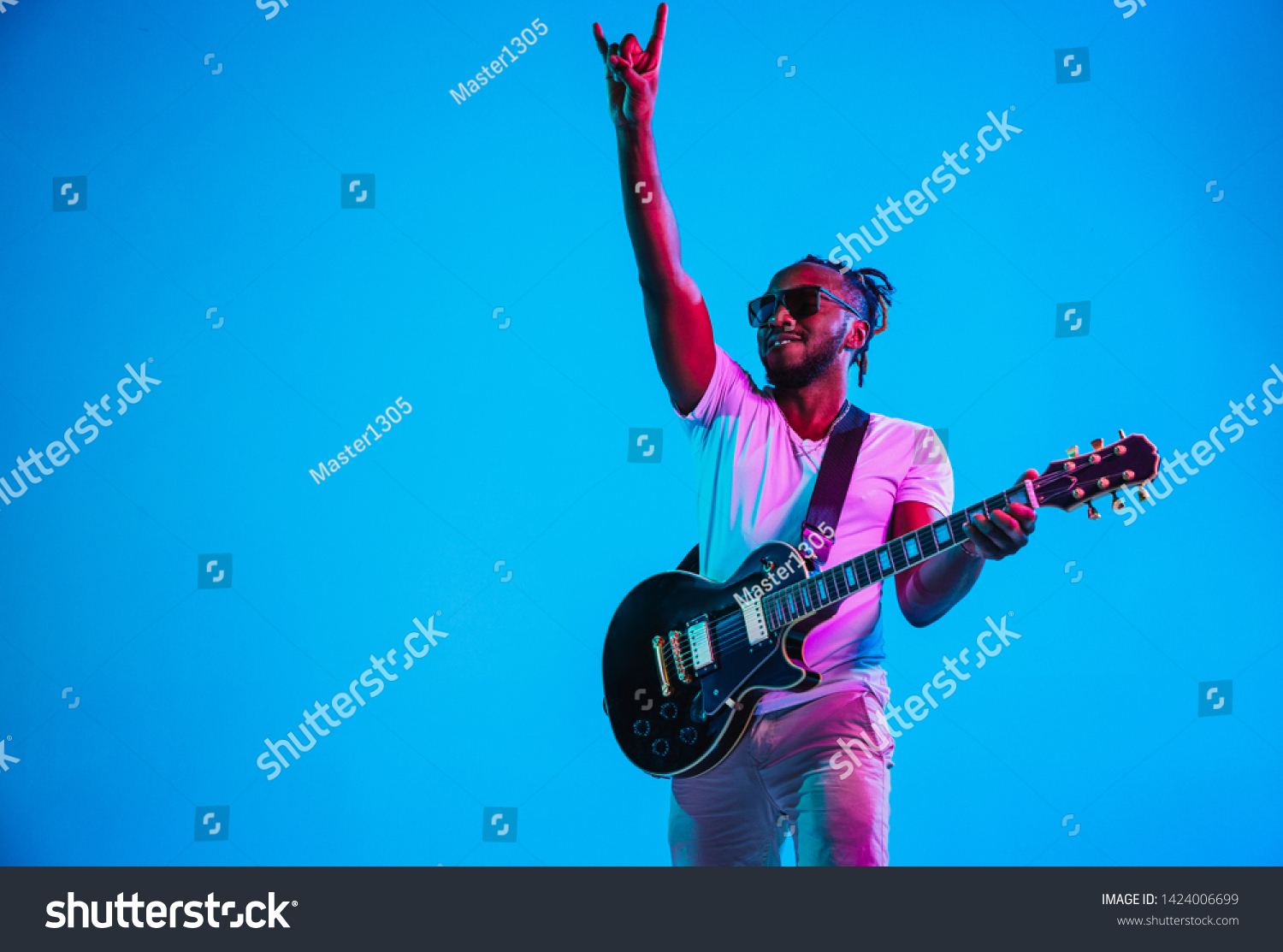 Young african-american musician playing the guitar like a rockstar on blue studio background in neon light. Concept of music, hobby. Joyful attractive guy improvising. Retro colorful portrait. #1424006699