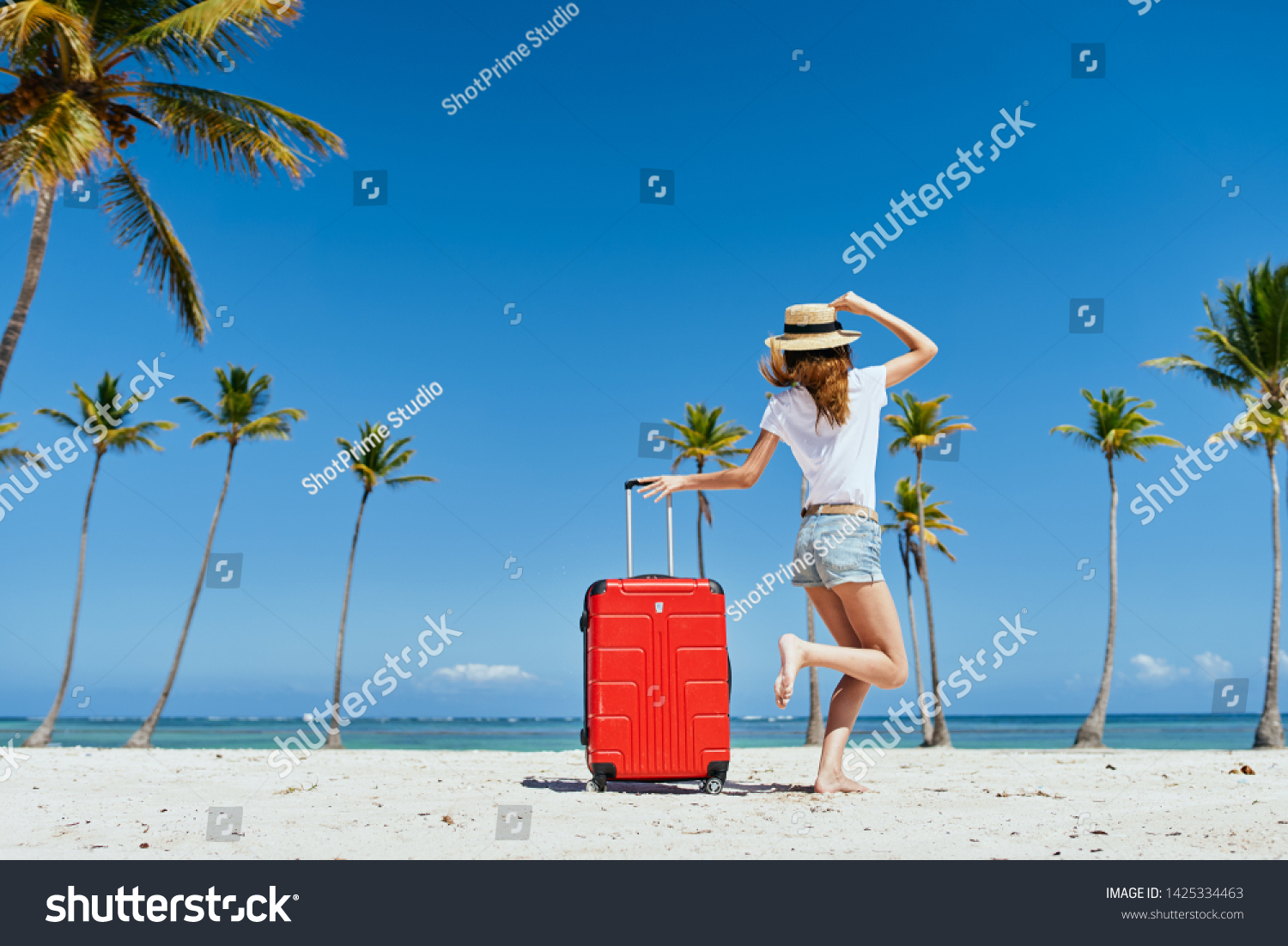 A woman in shorts and a t-shirt leaned on the handle of a suitcase and stands on the sand with a hat on her head                        #1425334463