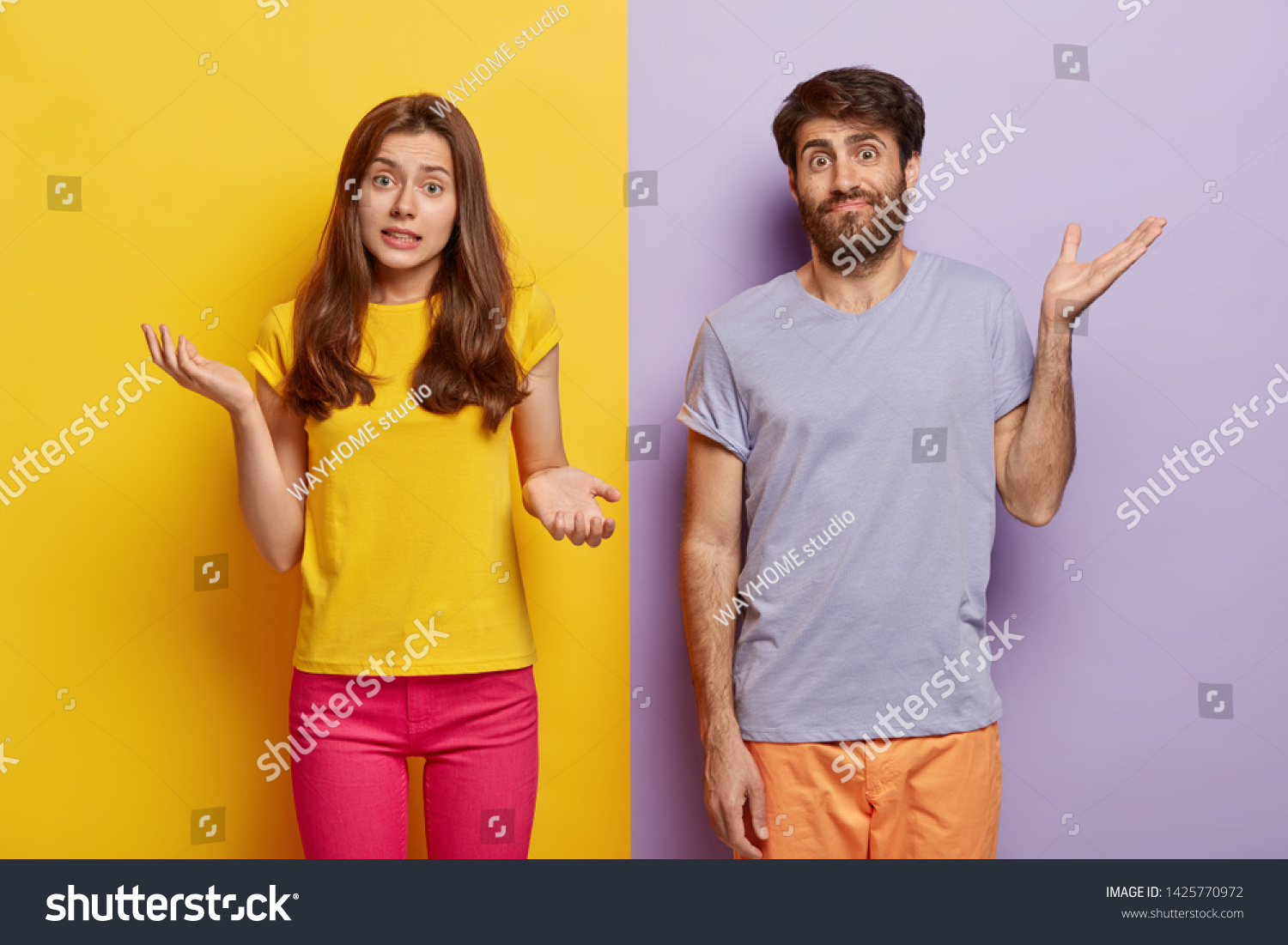 Indifferent unbothered woman and man spread hands sideways, have no idea, dressed in casual outfit, pose against different color background. Confused questioned couple with clueless expression indoor #1425770972