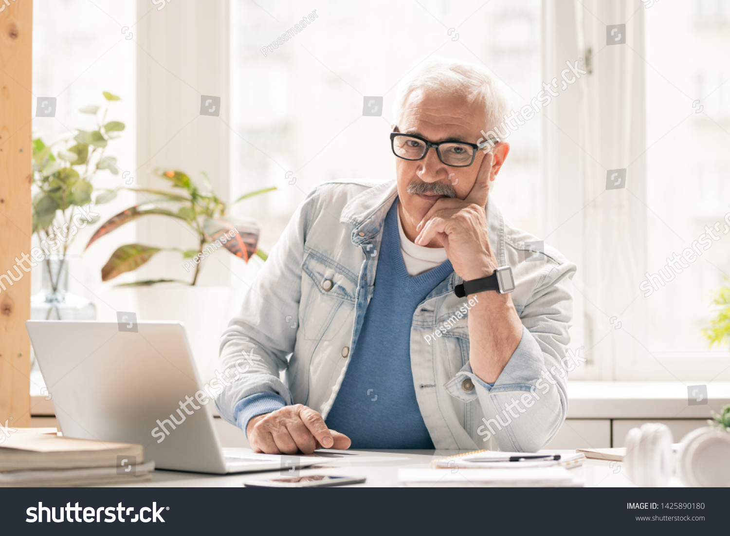 Mature pensive businessman in casualwear looking at you while sitting by desk in front of laptop in office #1425890180