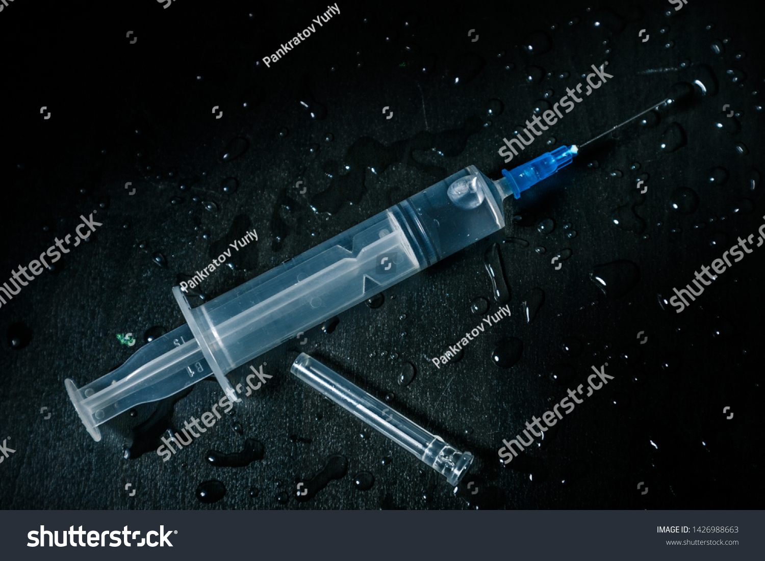 A syringe with a clear liquid lies on a dirty dark background covered with drops. #1426988663