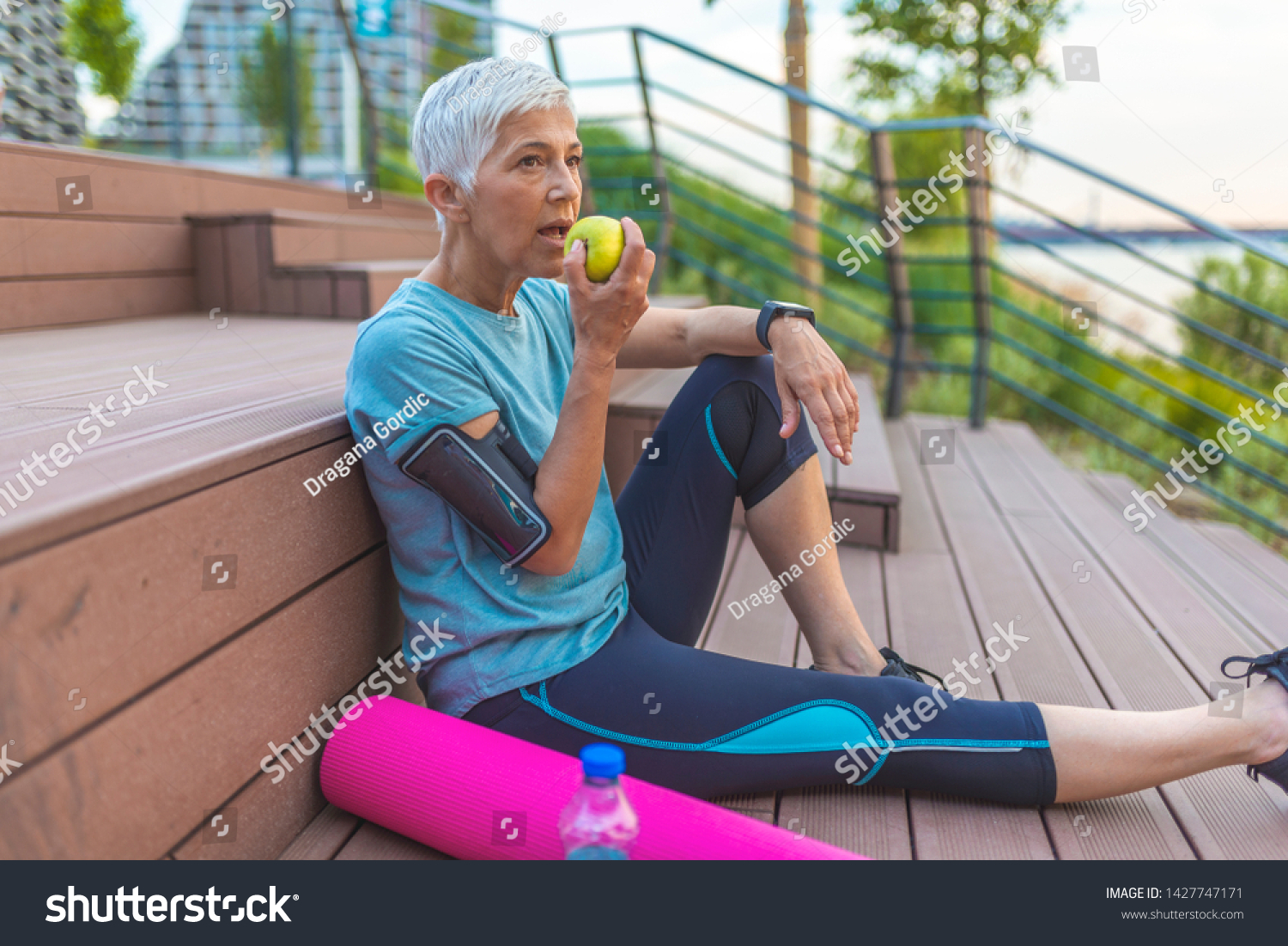 Senior woman with apple and water. Smiling woman eating green apple. Portrait of senior woman sitting on stairs after exercising. Sporty woman eating apple #1427747171