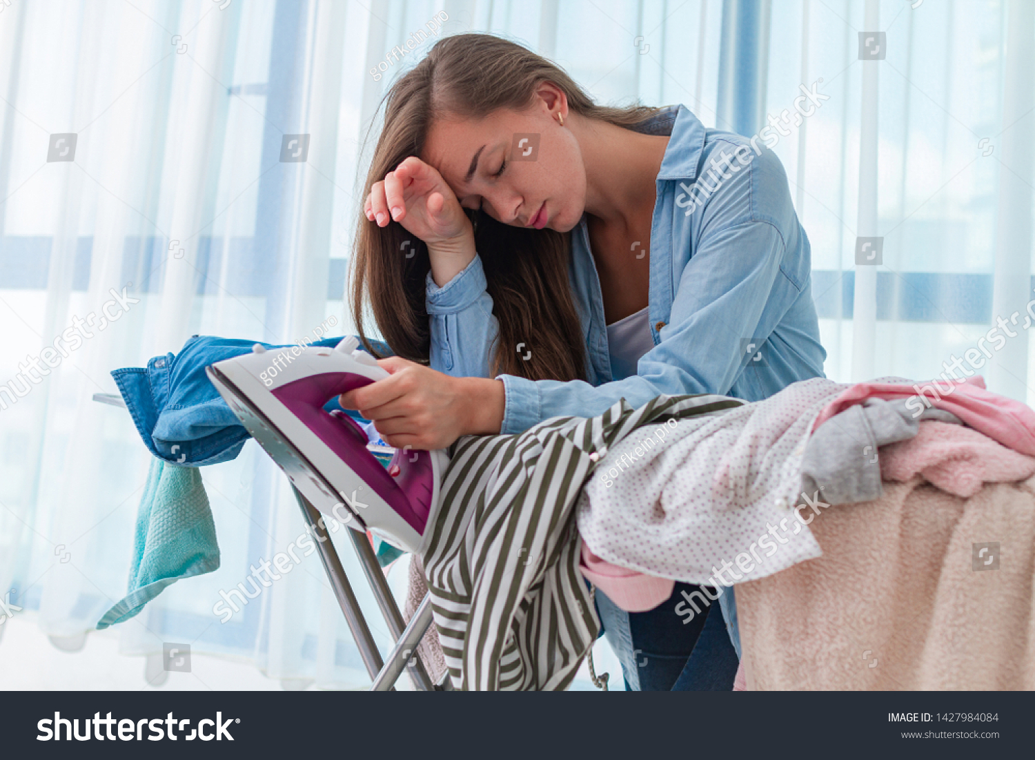 Ironing woman feeling tired from ironing pile of clothes after laundry at home. Household chores and routine #1427984084
