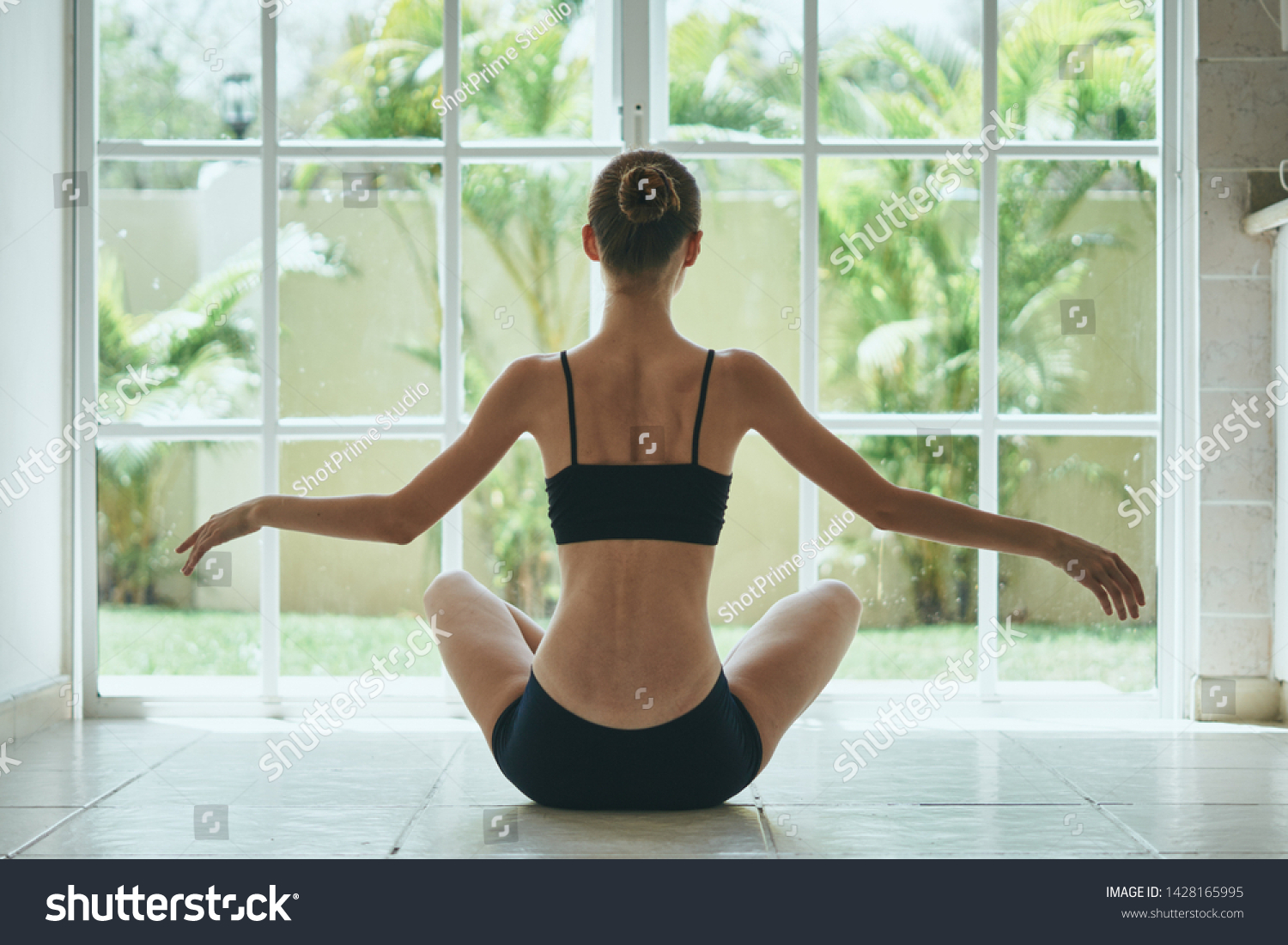 A woman in a short T-shirt and shorts sits in front of the window indoors and meditates                           #1428165995