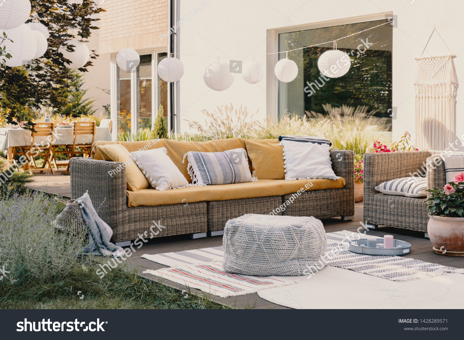 Pouf next to rattan couch and armchair on wooden terrace with flowers and lamps #1428289571
