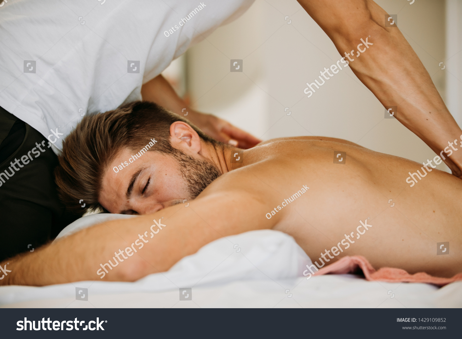 Male to male massage from young Asian therapist giving massage to muscle gym fit Caucasian man who laying on the bed in massage room. Remedial oil massage is performing on client. #1429109852