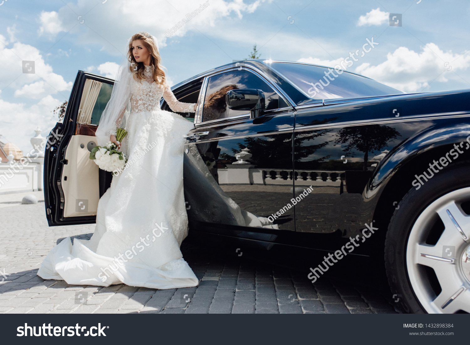 Stylish Bride with Flower Bouquet near Limousine. Pretty Young Woman Dressed White Wedding Dress and Jewelry Diadem with Veil. Girl Holding Roses Bunch and Standing near Black Car. Outside Photo #1432898384