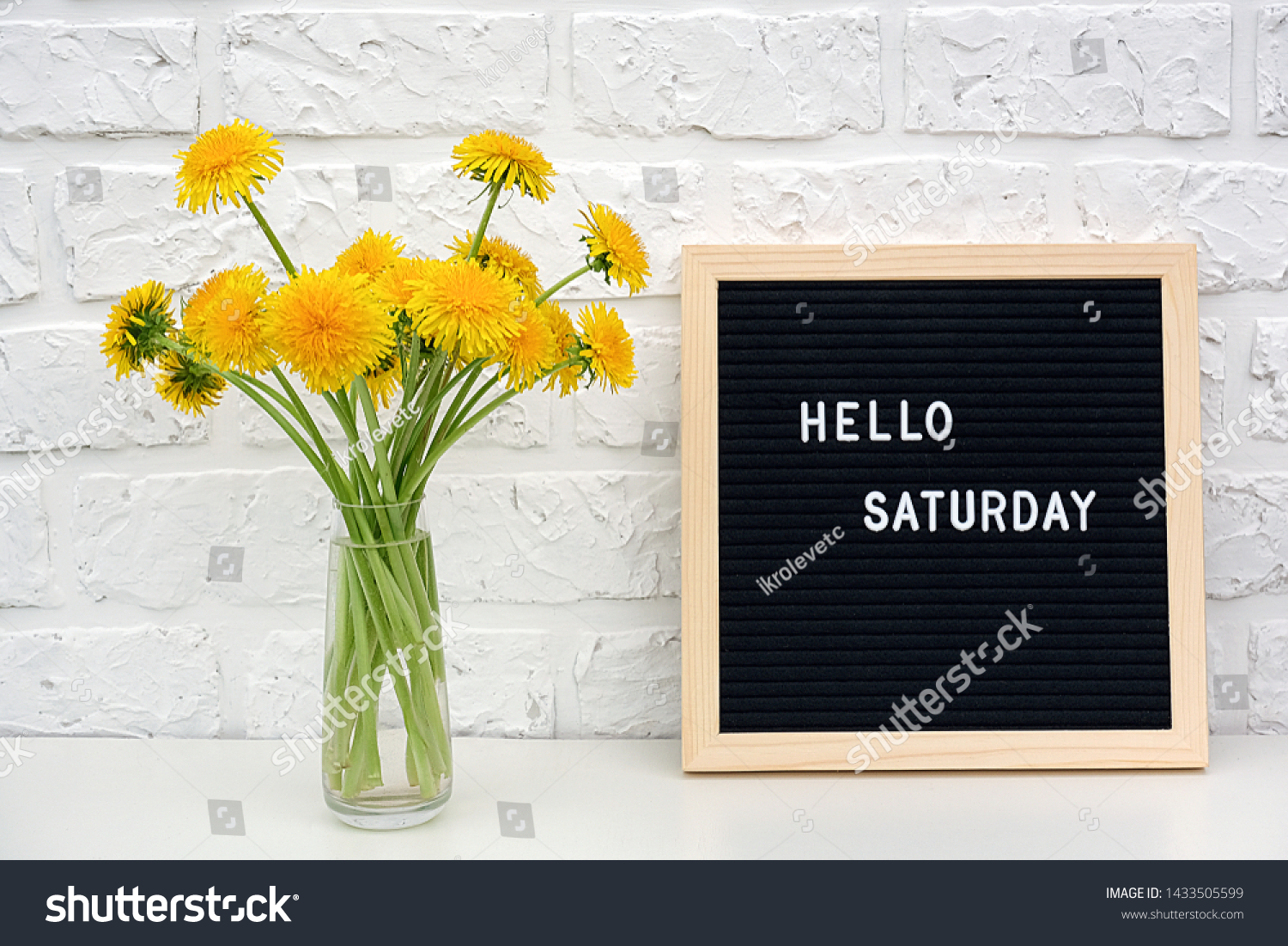 Hello Saturday words on black letter board and bouquet of yellow dandelions flowers on table against white brick wall. Concept Happy Saturday. Template for postcard. #1433505599