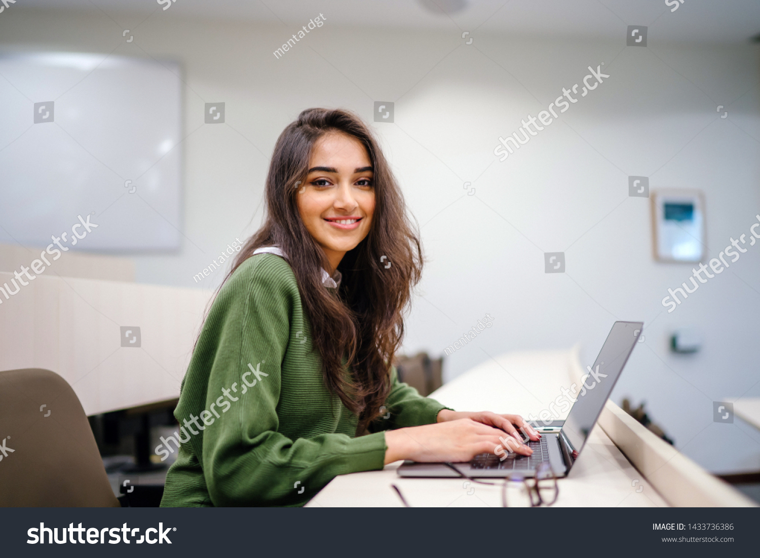 Portrait of a beautiful, young and intelligent-looking Indian Asian woman student wearing a white shirt and green tracker smiling as she works on her laptop in a university classroom.  #1433736386