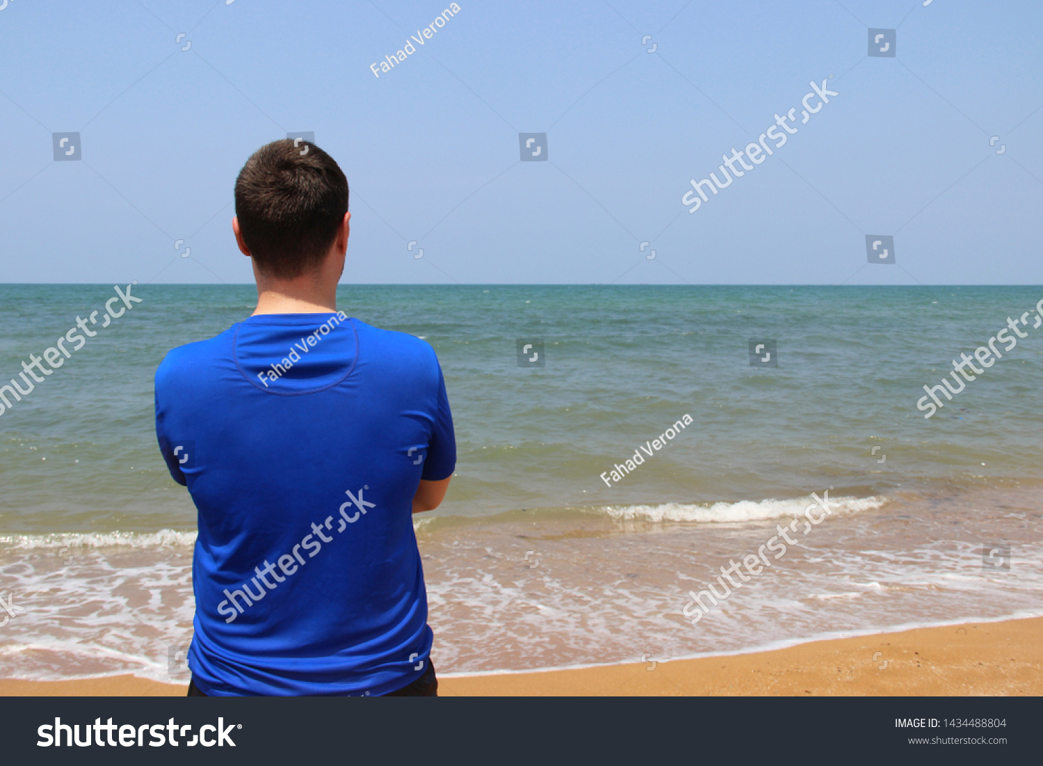a man stands with his back in a blue t-shirt for swimming against the background of the sea and sand. sunny summer day. the man has dark hair #1434488804