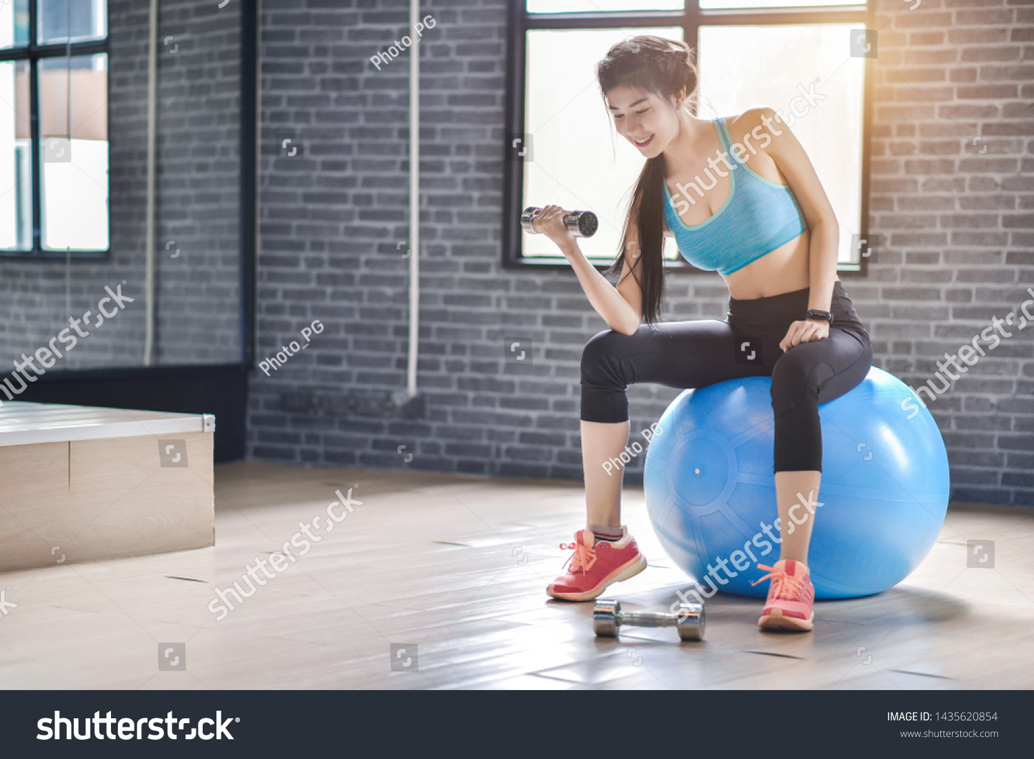 Beautiful fitness woman lifting dumbbell in the morning with ball to ell-developed muscles by strength training in gym #1435620854
