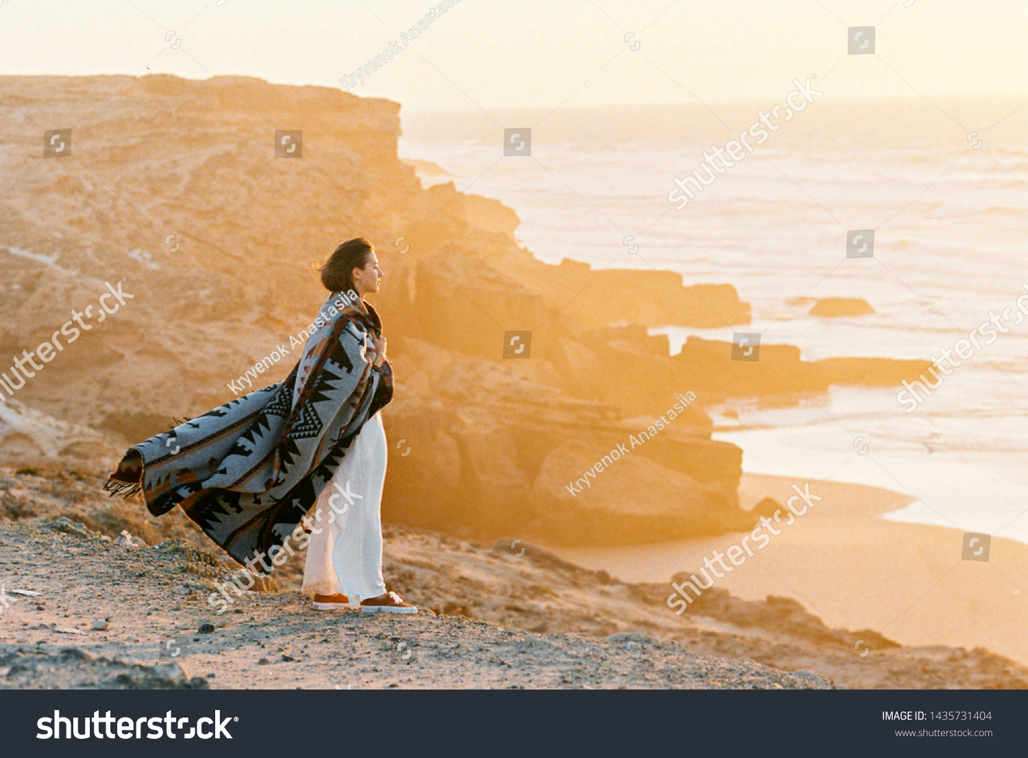 Young woman with flying blanket standing on cliff's edge and looking to the ocean on sunset. Wanderlust concept. #1435731404