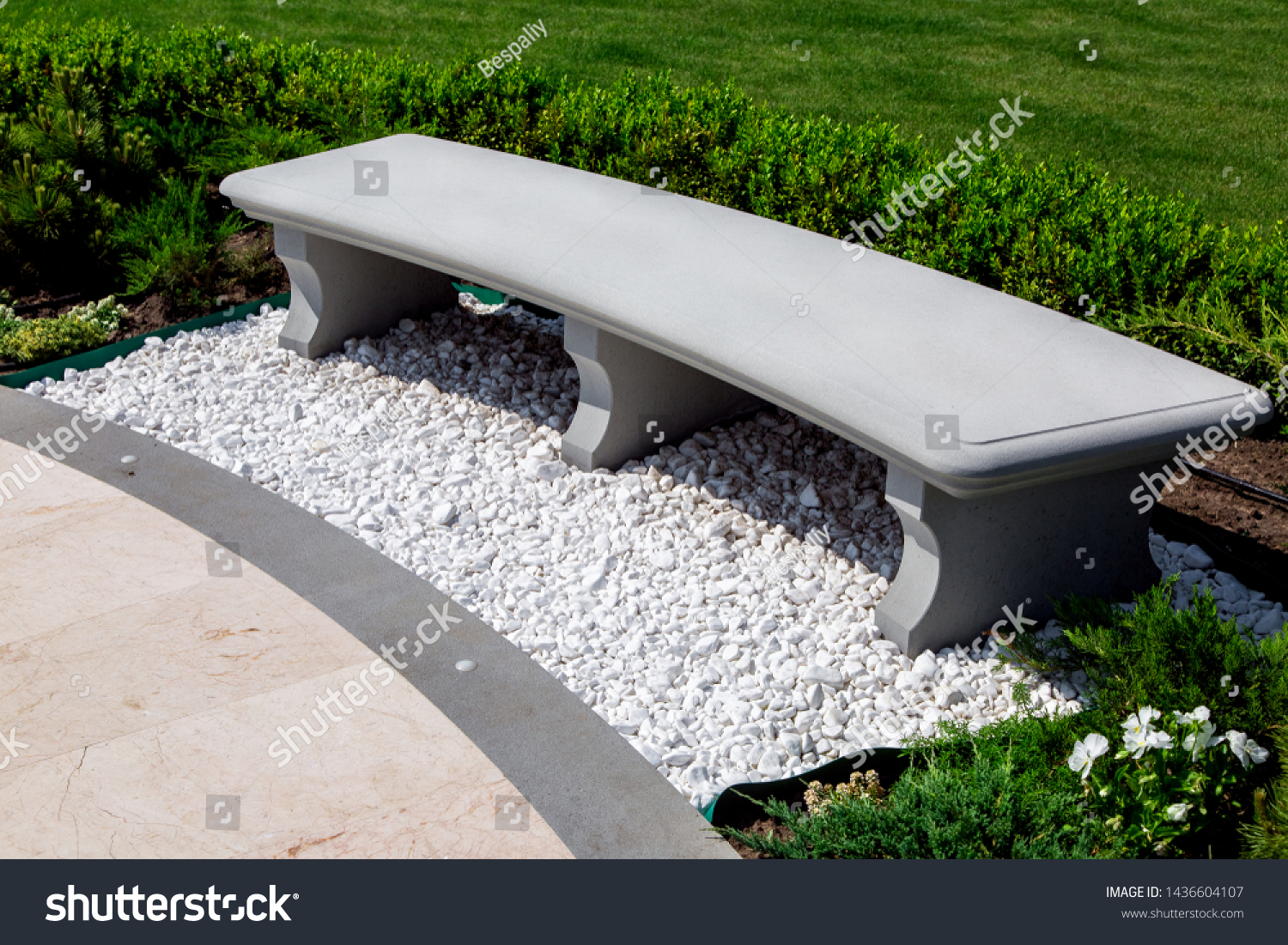 stone gray bench strewn with white stone pebbles in a garden with boxwood bushes and a green lawn with plants, close up of a place for sitting and resting near a marble tile walkway on a sunny summer. #1436604107