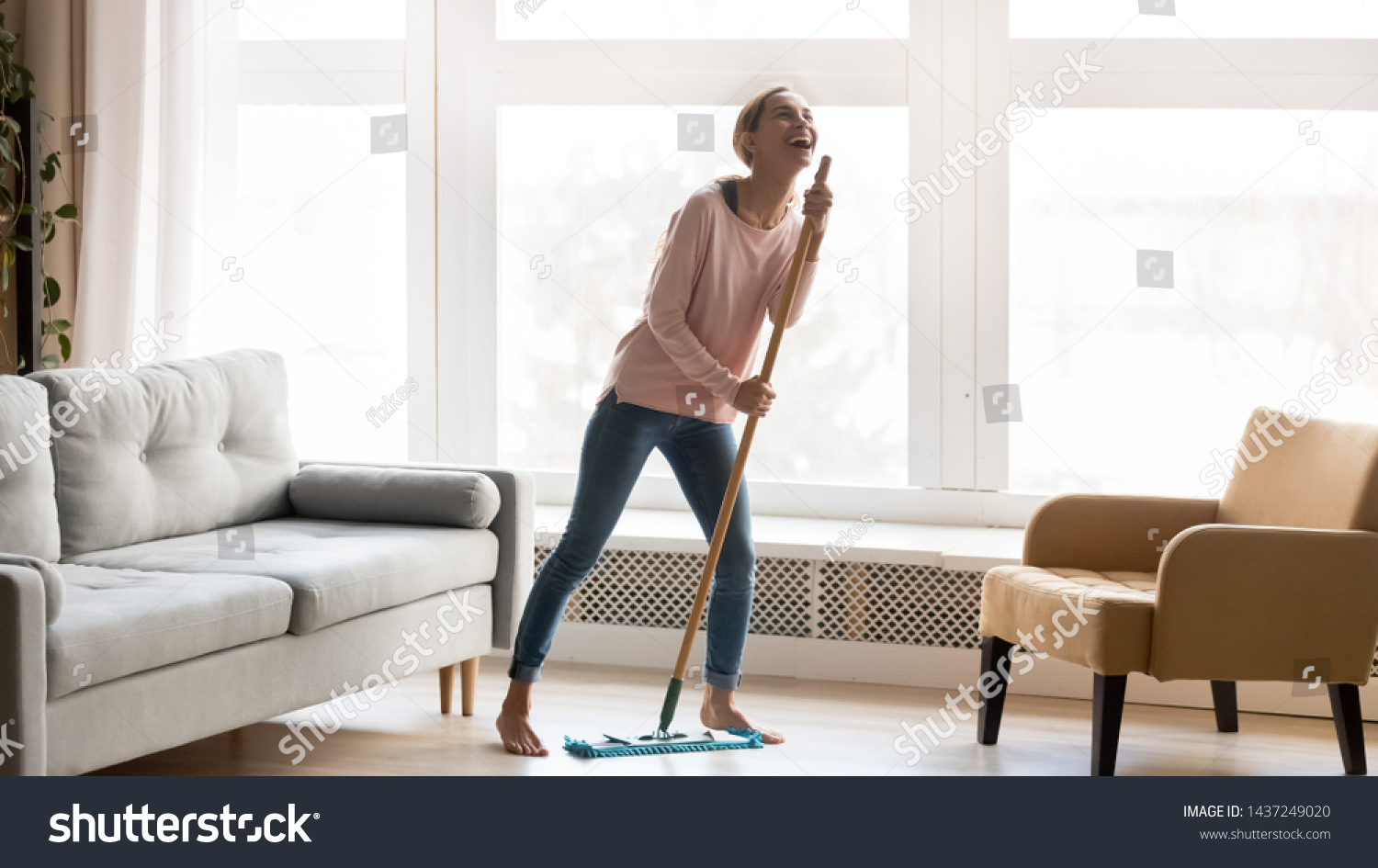 Carefree happy young woman cleaning house living room have fun dancing with mop, smiling overjoyed millennial girl feel excited enjoy making home chores sing entertain using floor broom or Swiffer #1437249020