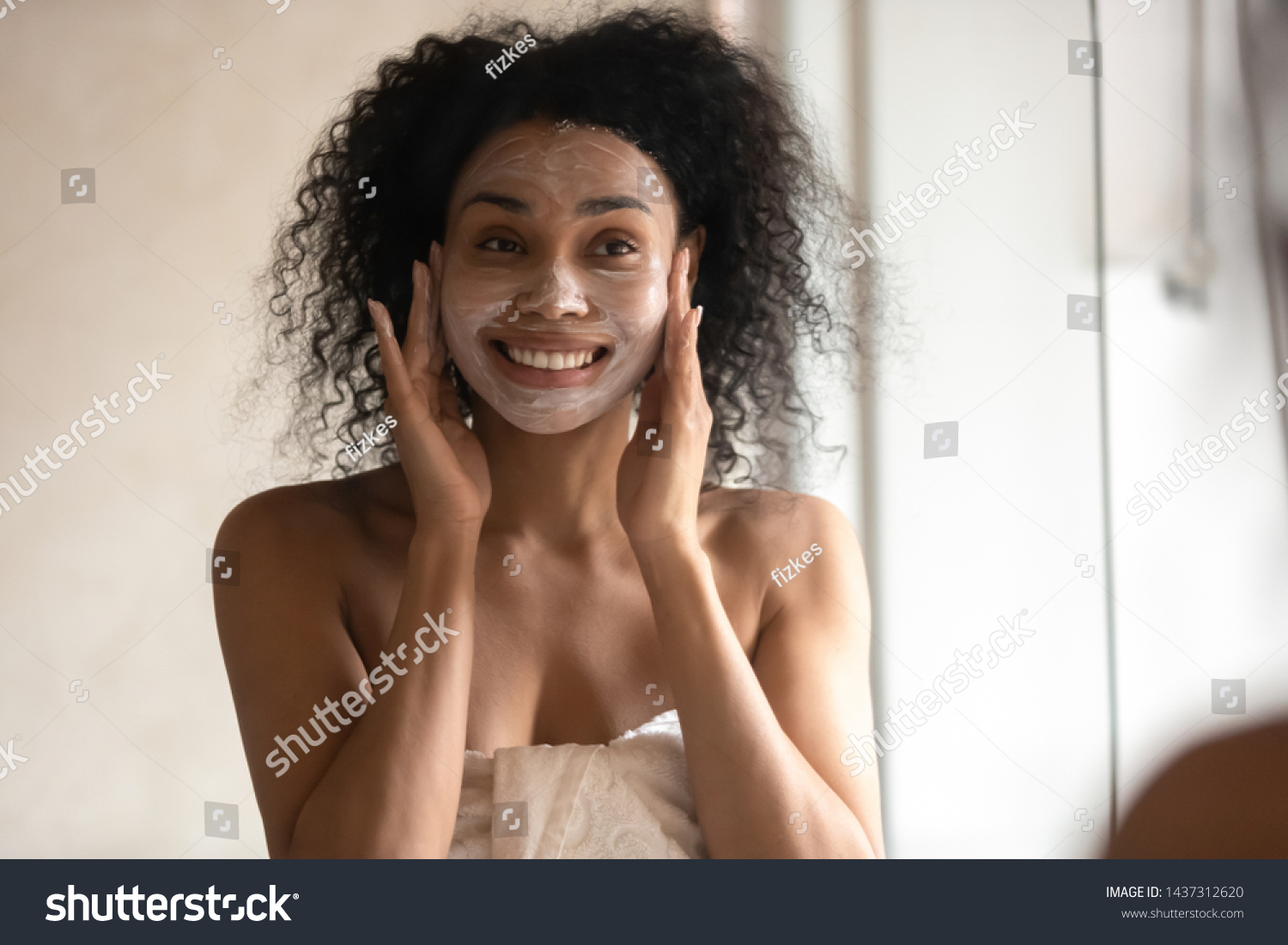 African 30s young woman after shower is wrapped in towel looking in mirror pampering herself applying face mask use anti-aging beauty product feeling happy, skin care, everyday morning routine concept #1437312620