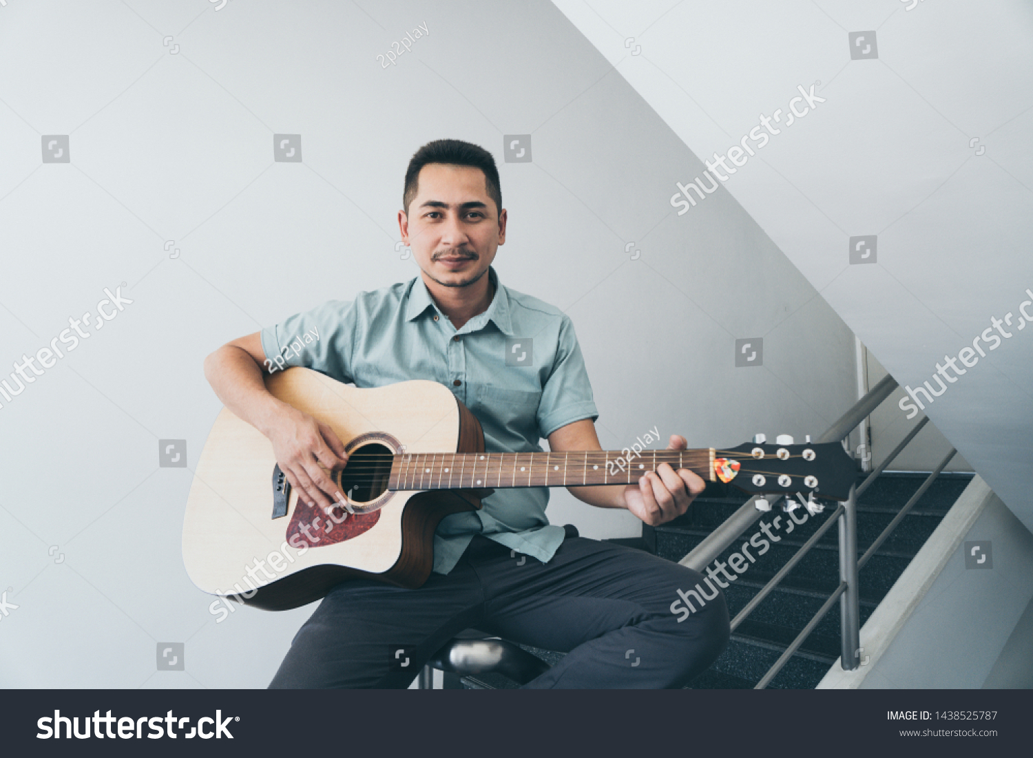 Cheerful guitarist. Cheerful handsome young man playing guitar and smiling while sitting on banister, process color and effect grain. #1438525787