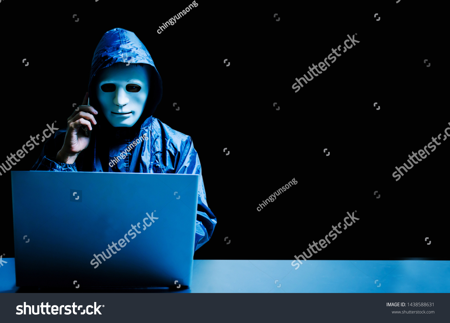 Anonymous computer hacker in white mask and hoodie. Obscured dark face using laptop computer for cyber attack and calling on cellphone, Data thief, internet attack, darknet and cyber security concept. #1438588631