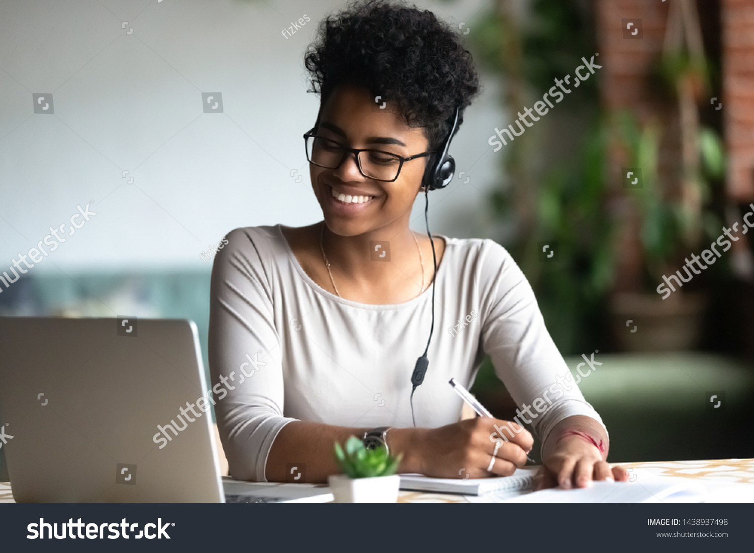 Smiling african American millennial female student in headphones and glasses sit at desk watch webinar making notes, happy biracial young woman in earphones work study using computer write in notebook #1438937498