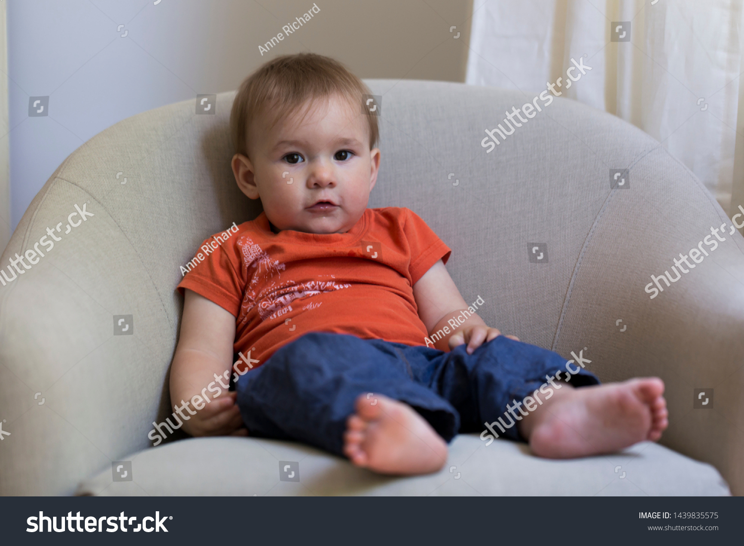 Horizontal full length portrait of cute fair toddler girl in orange shirt and blue pants lounging in armchair with adorable innocent expression #1439835575