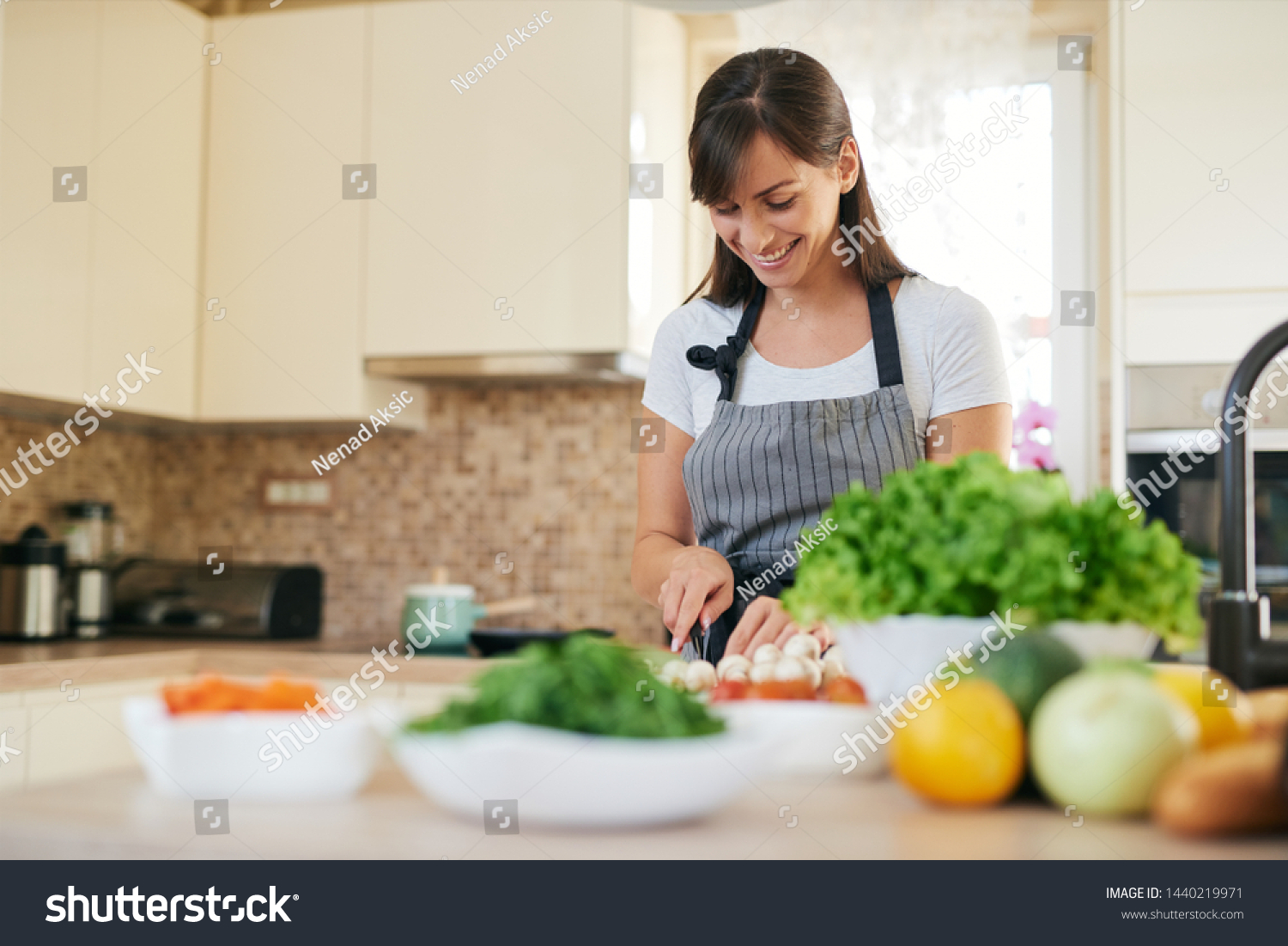 Beautiful smiling dedicated Caucasian brunette in apron standing in kitchen and chopping mushrooms. On table are lots of vegetables. Cooking at home concept. #1440219971
