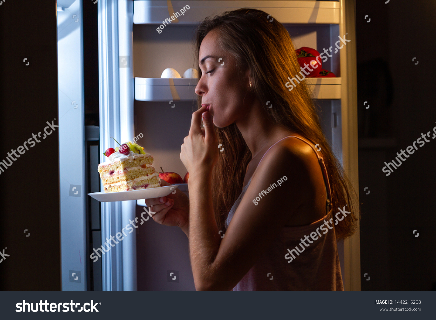 Hungry woman in pajamas enjoys sweet cake at night near refrigerator. Stop diet and gain extra pounds due to high carbs food and unhealthy eating #1442215208