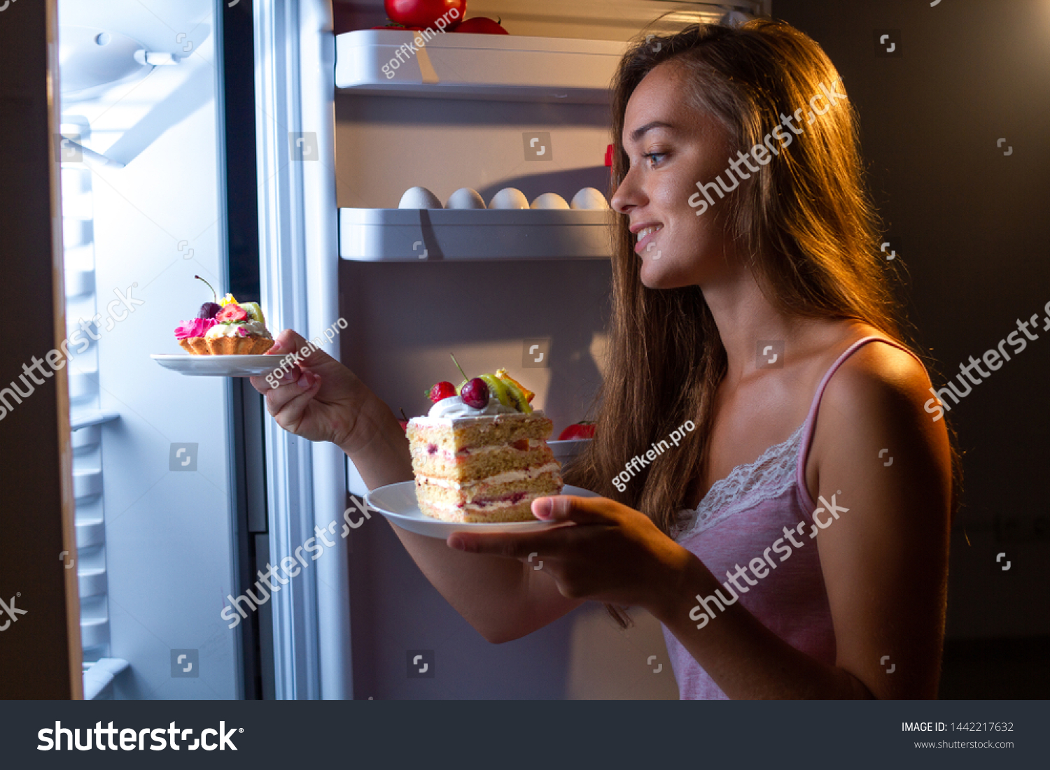 Hungry woman in pajamas eats and enjoys cakes  at night near refrigerator. Stop diet and gain extra pounds due to carbs food and unhealthy night eating  #1442217632