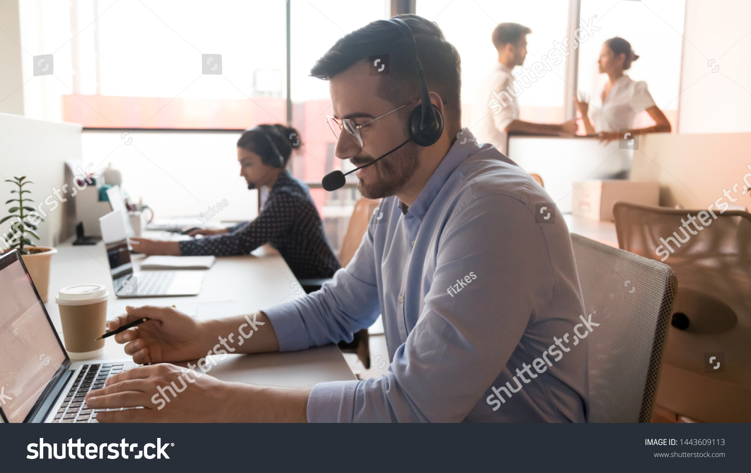 Male professional call center telesales agent wear wireless headset using computer in customer care support service office with team, businessman operator telemarketer working on laptop at workplace #1443609113