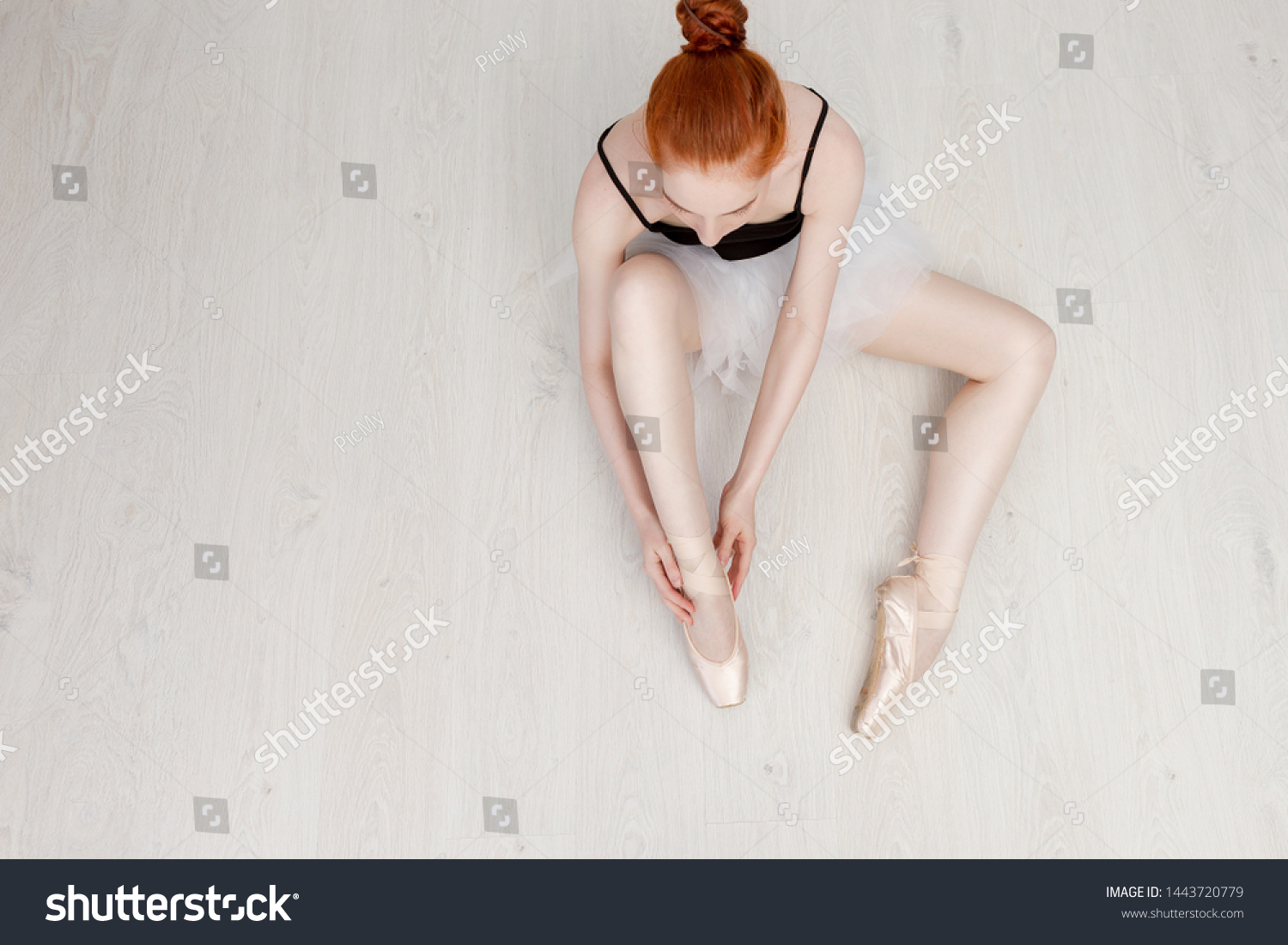 Nice ballerina sits on the white floor and dresses a beige pointe shoe in the studio. She wears a light dance wear and a peach tutu. Top view photo. Horizontal. Copy space #1443720779