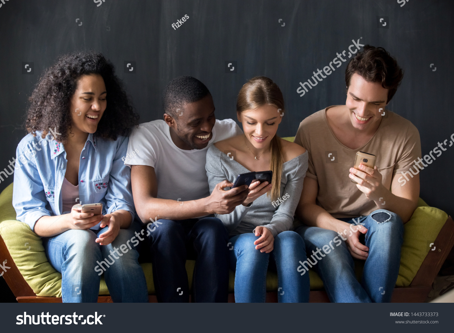 Happy excited smiling diverse friends sitting on couch, discussing new apps for mobile phones, sharing contacts, laughing at funny videos or party photos, playing games, teen mobile addiction concept. #1443733373