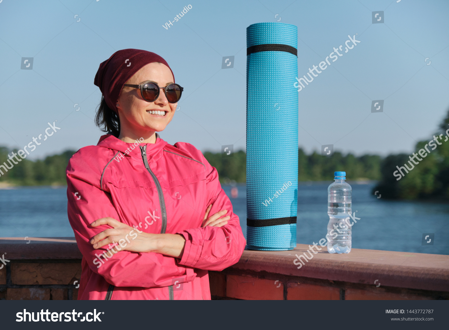 Middle-aged confident sports woman with yoga mat and bottle of water with crossed folded arms, an outdoor evening sunny park near the river. Active healthy lifestyle of an age female #1443772787