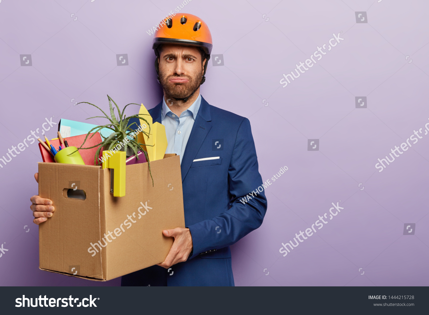Fatigue male worker carries heavy cardboard box with office stuff, moves in new cabinet, wears helmet and suit, has dissatisfied tired facial expression, busy carrying paper container, stands indoor #1444215728