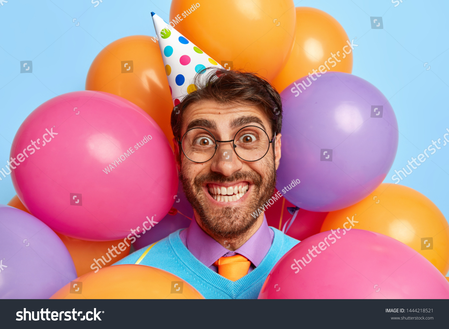 Headshot of handsome joyful birthday man celebrates anniversary in family circle, has perfect festive mood, receives congratulations and presents, wears cone paper hat, makes photo with balloons #1444218521