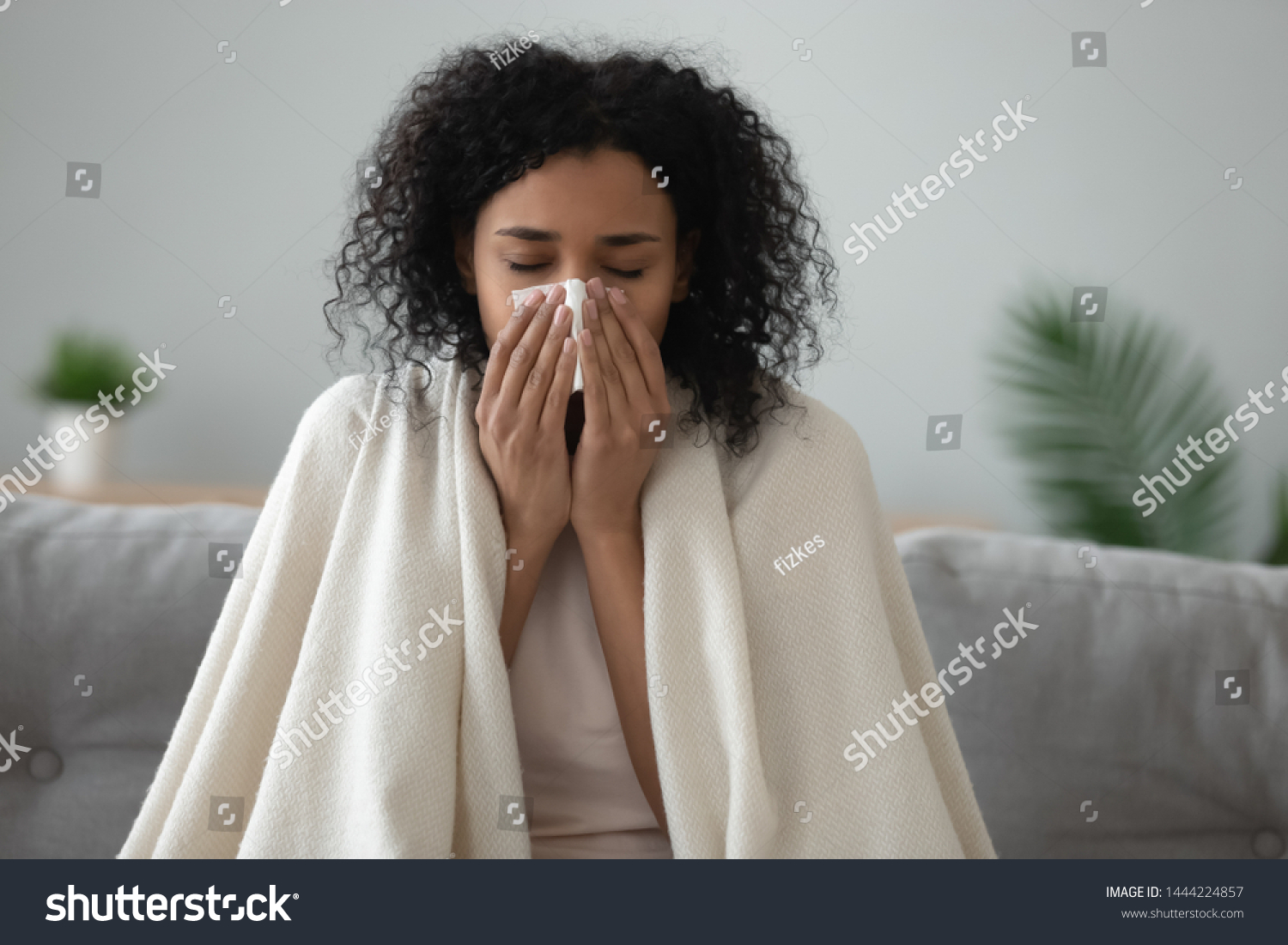 Ill african young woman covered with blanket blowing running nose got fever caught cold sneezing in tissue sit on sofa, sick allergic black girl having allergy symptoms coughing at home, flu concept #1444224857