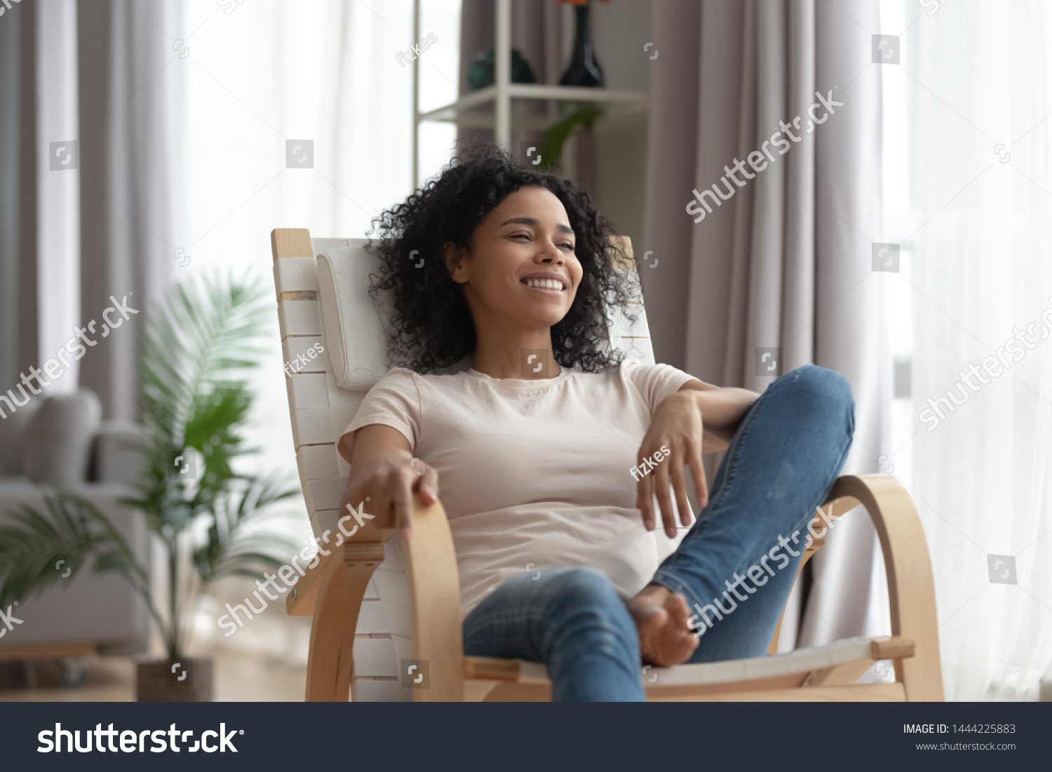 Smiling calm young black woman relaxing on comfortable wooden rocking chair in living room, happy healthy black girl enjoy breathing fresh air resting in armchair at home feel stress free on weekend #1444225883