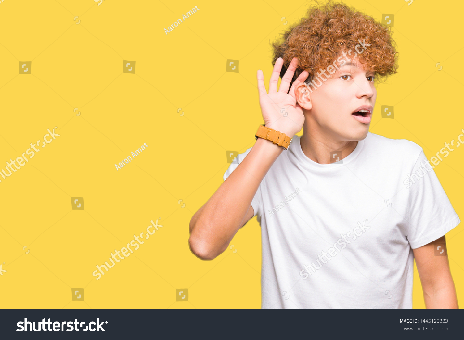Young handsome man with afro hair wearing casual white t-shirt smiling with hand over ear listening an hearing to rumor or gossip. Deafness concept. #1445123333