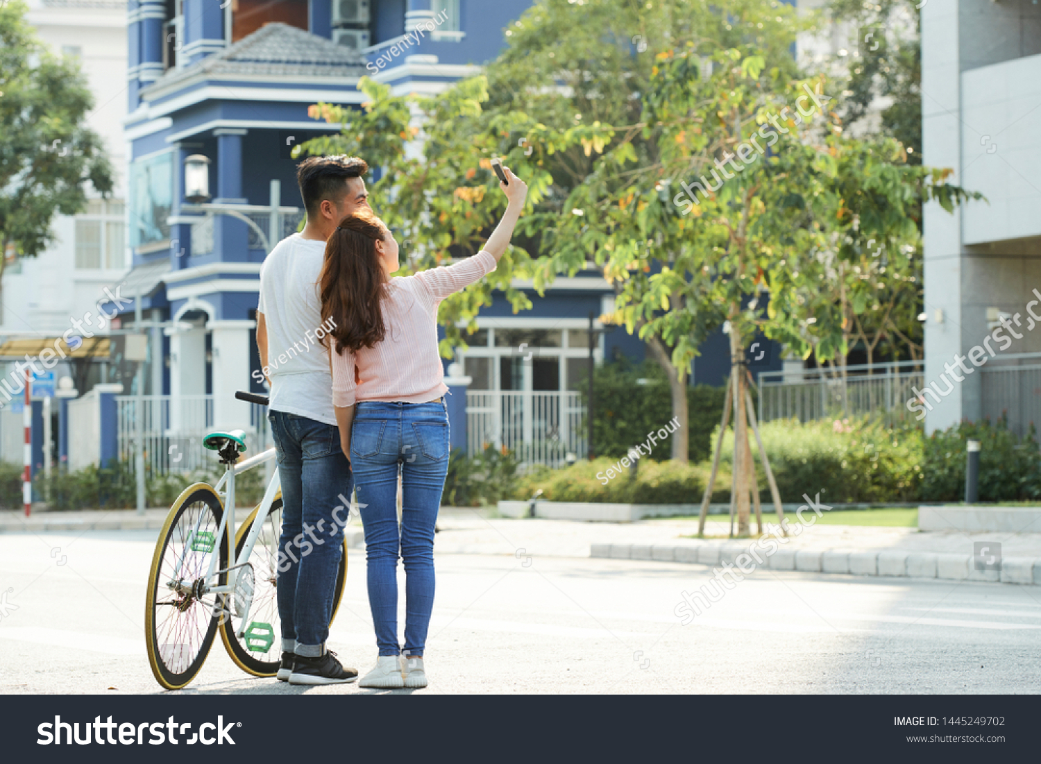 Rear view of Asian young couple standing with bicycle on the street and making selfie portrait on mobile phone #1445249702