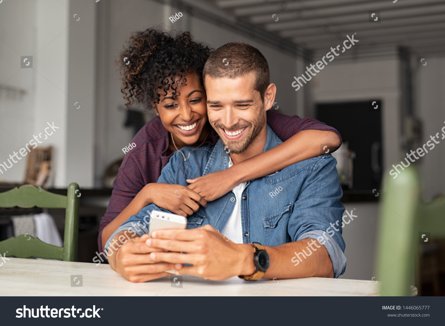 Smiling young couple embracing while looking at smartphone. Multiethnic couple sharing social media on smart phone. Smiling african girl embracing from behind her happy boyfriend while using cellphone #1446065777