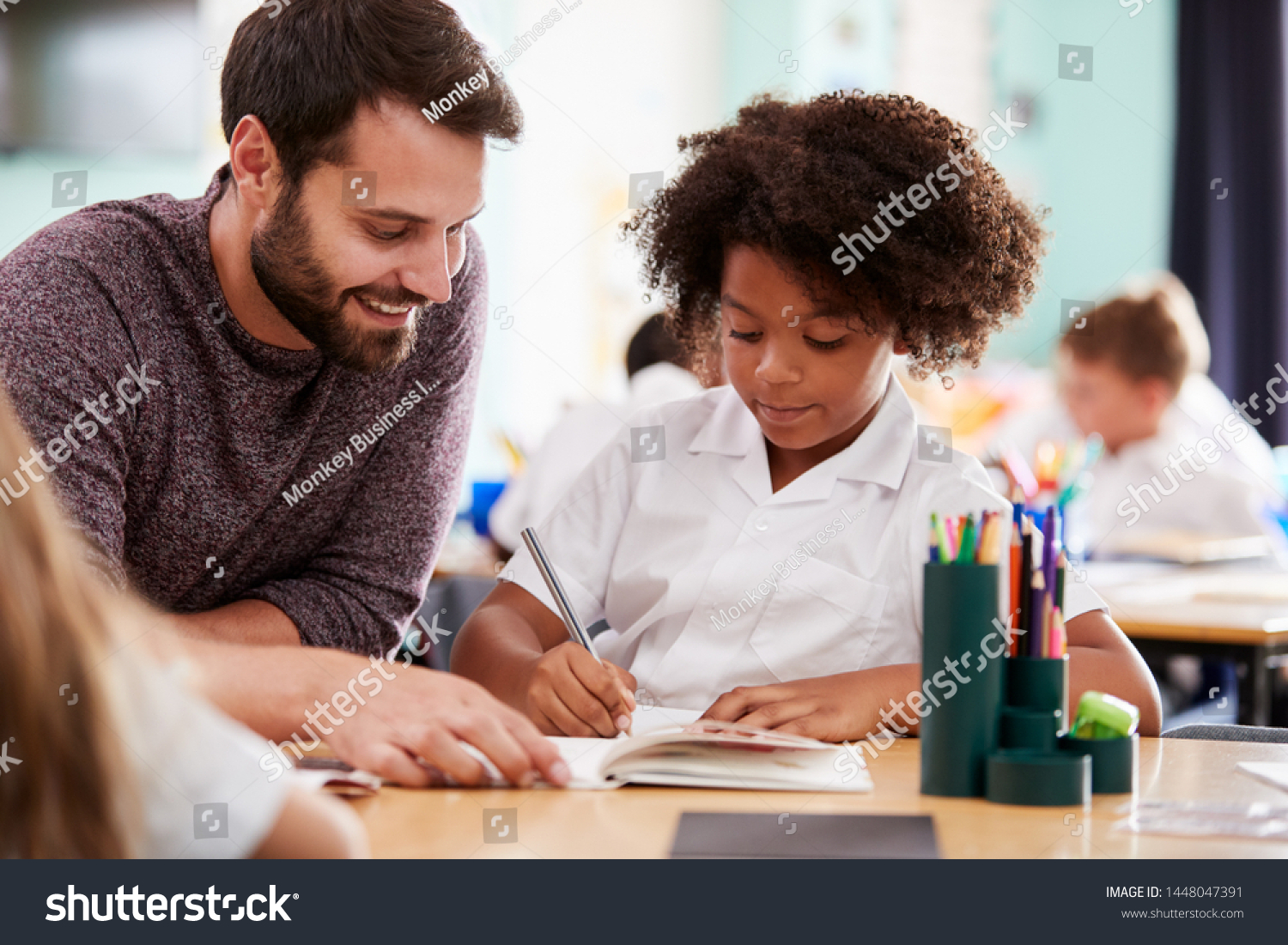 Male Elementary School Teacher Giving Female Pupil Wearing Uniform One To One Support In Classroom #1448047391