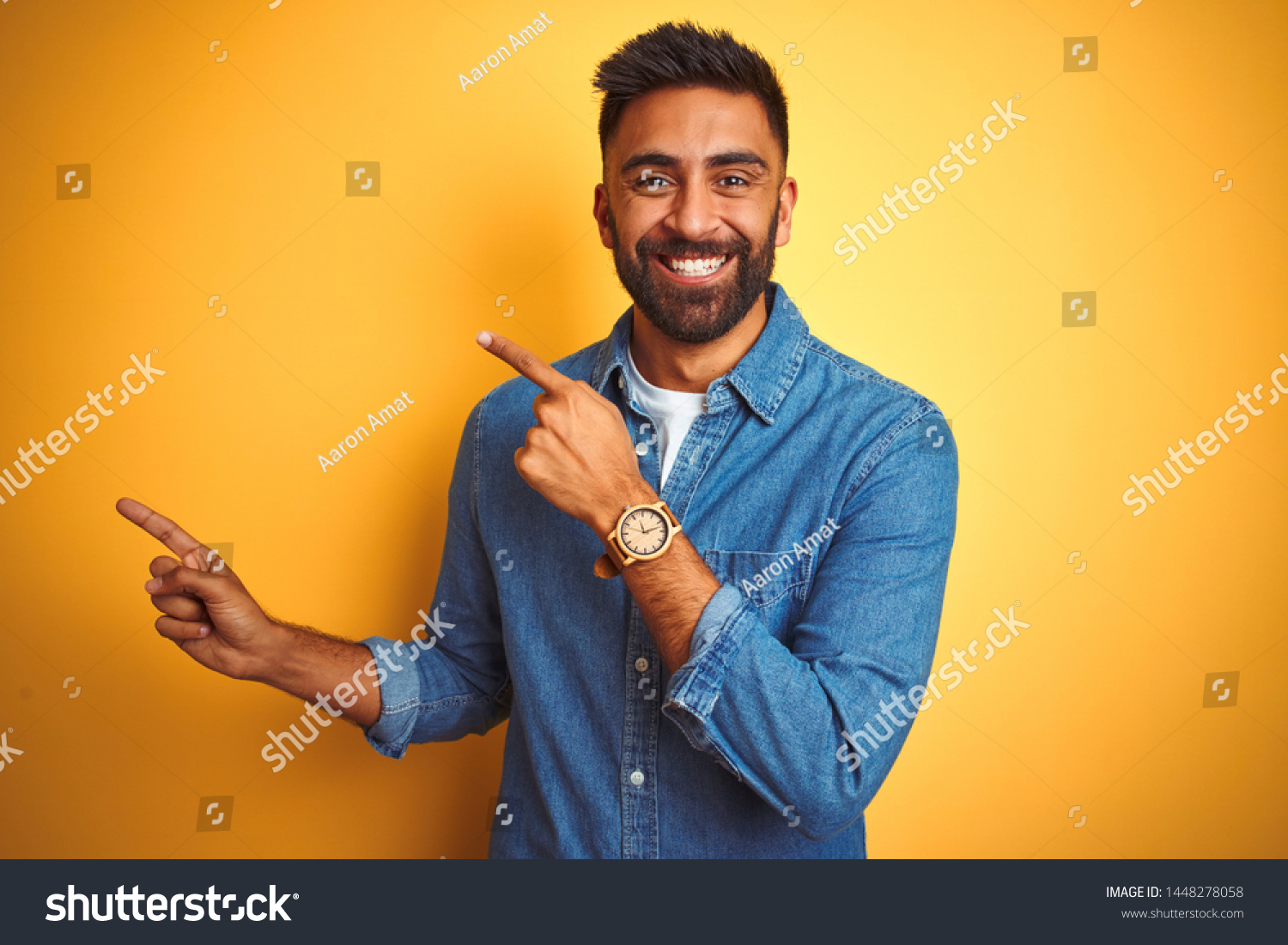 Young indian man wearing denim shirt standing over isolated yellow background smiling and looking at the camera pointing with two hands and fingers to the side. #1448278058